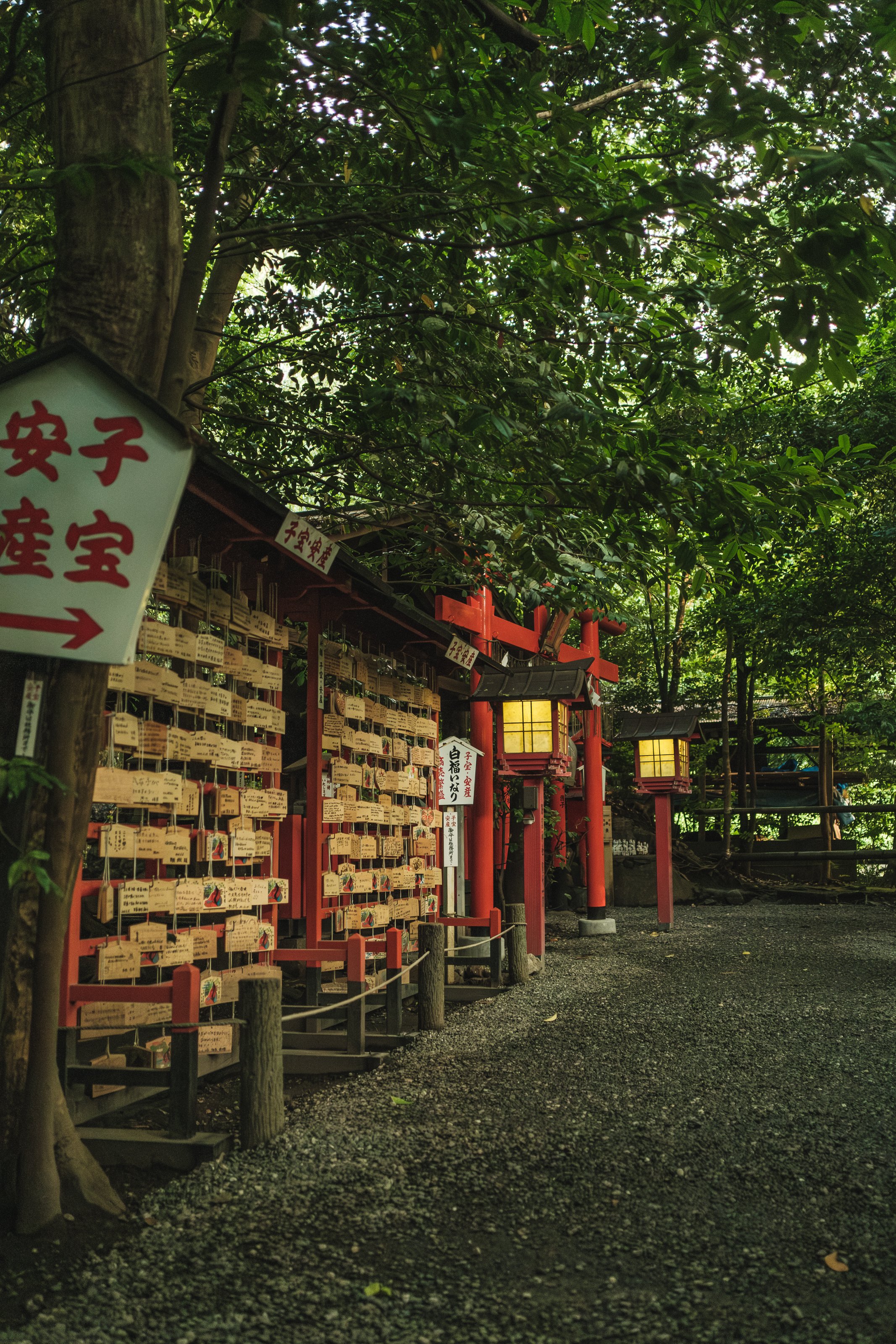 A Japanese shrine with red torii gates and lanterns surrounded by green trees, and wooden plaques hanging on a wall.