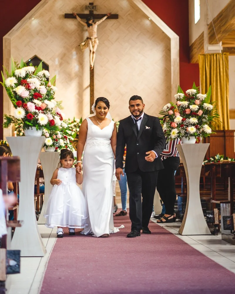 A bride and groom walking down the aisle of a church, accompanied by a young girl, with large flower arrangements on either side and a crucifix hanging on the wall behind them.