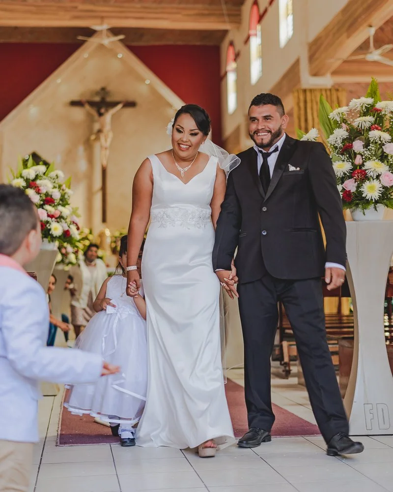A bride and groom walking down the aisle in a church, holding hands, smiling. The bride wears a white wedding gown and the groom a black suit. There are floral arrangements on either side and a large crucifix hanging on the wall behind them.