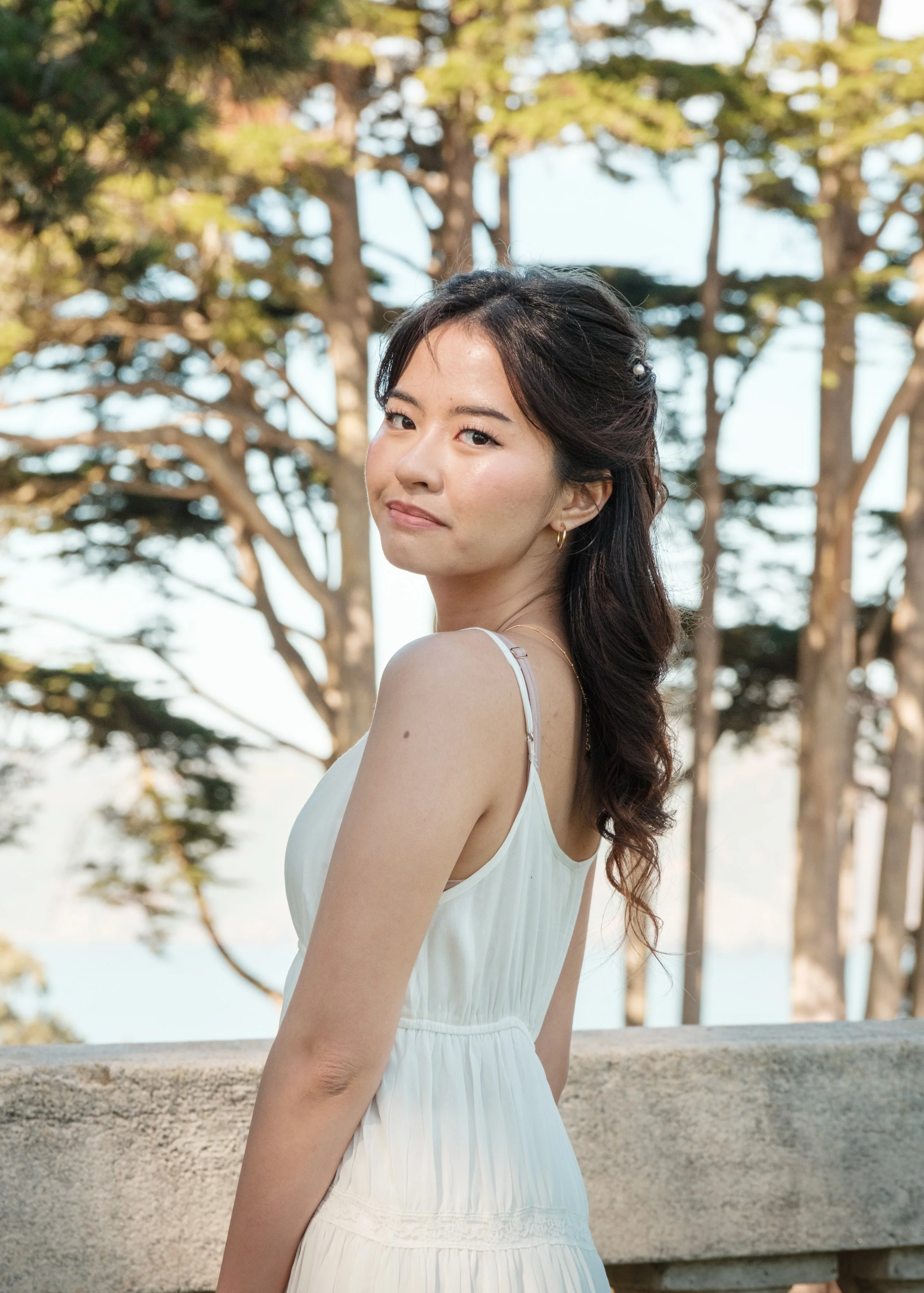 A young woman with dark hair and light skin, wearing a white sleeveless dress, standing outdoors near a stone railing with trees in the background.