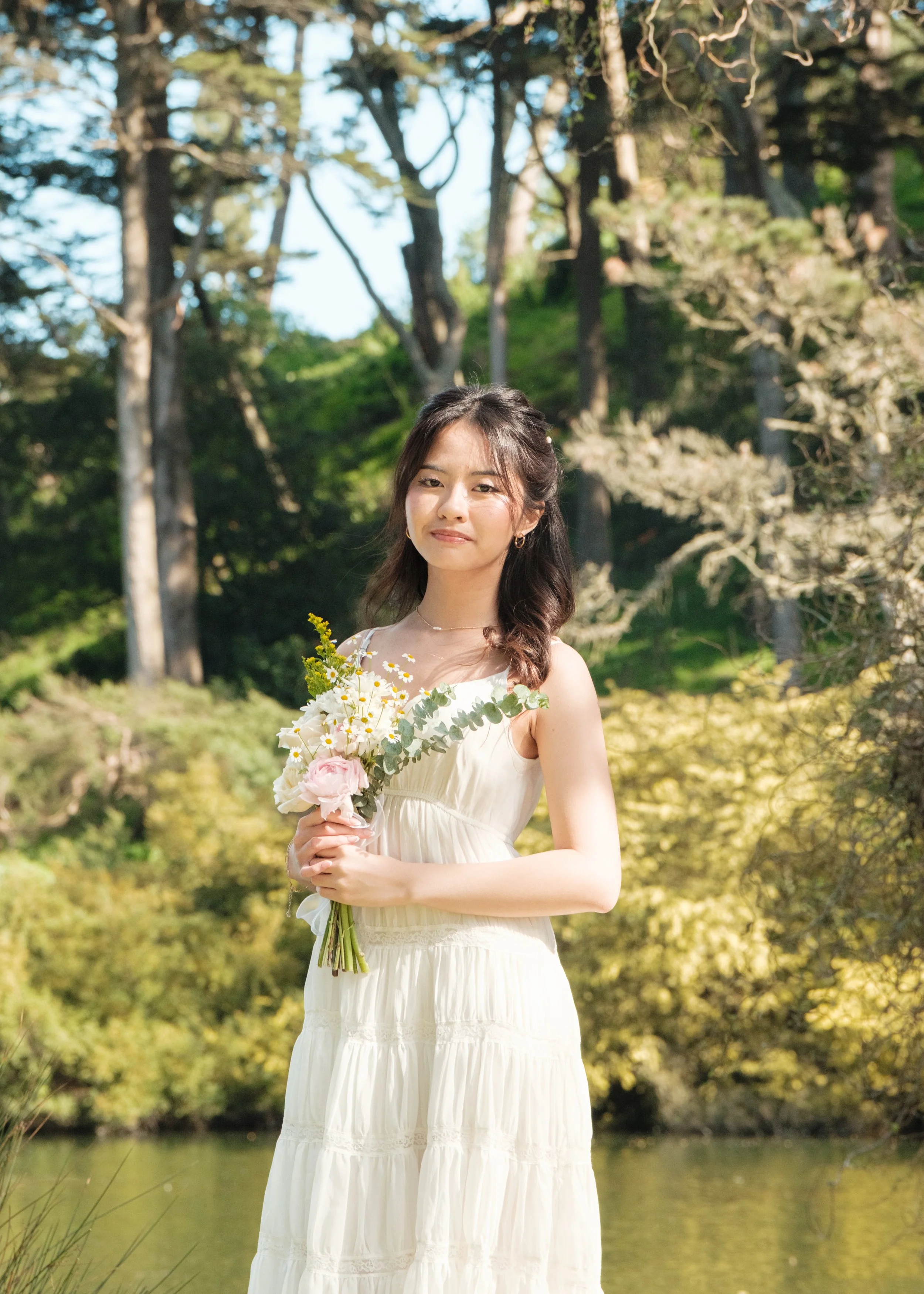 A woman wearing a white dress holding a bouquet of flowers outdoors with trees and greenery in the background.