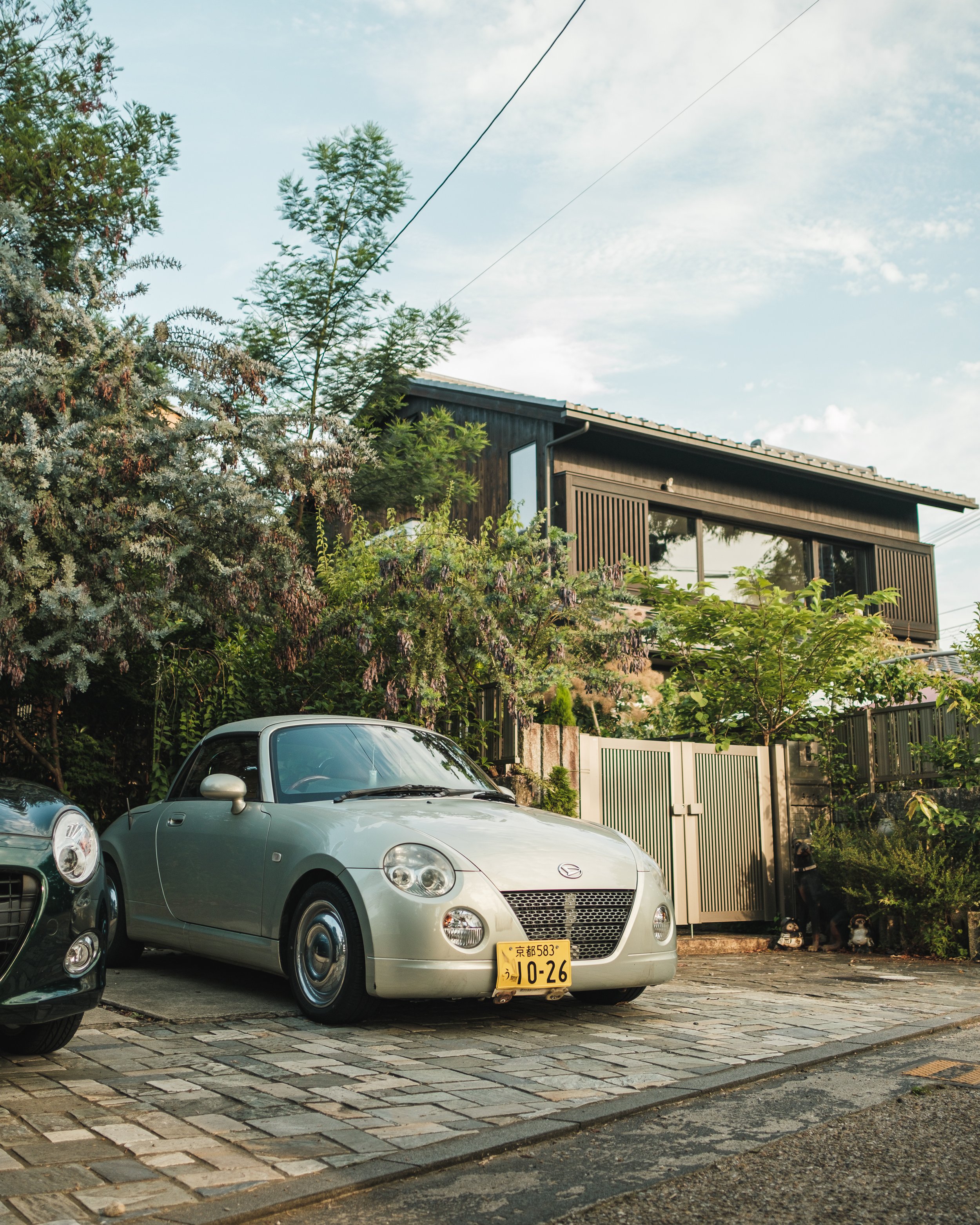 A silver Mazda MX-5 Miata convertible car parked on a stone-paved driveway in front of a modern house with large windows and surrounded by trees and greenery.