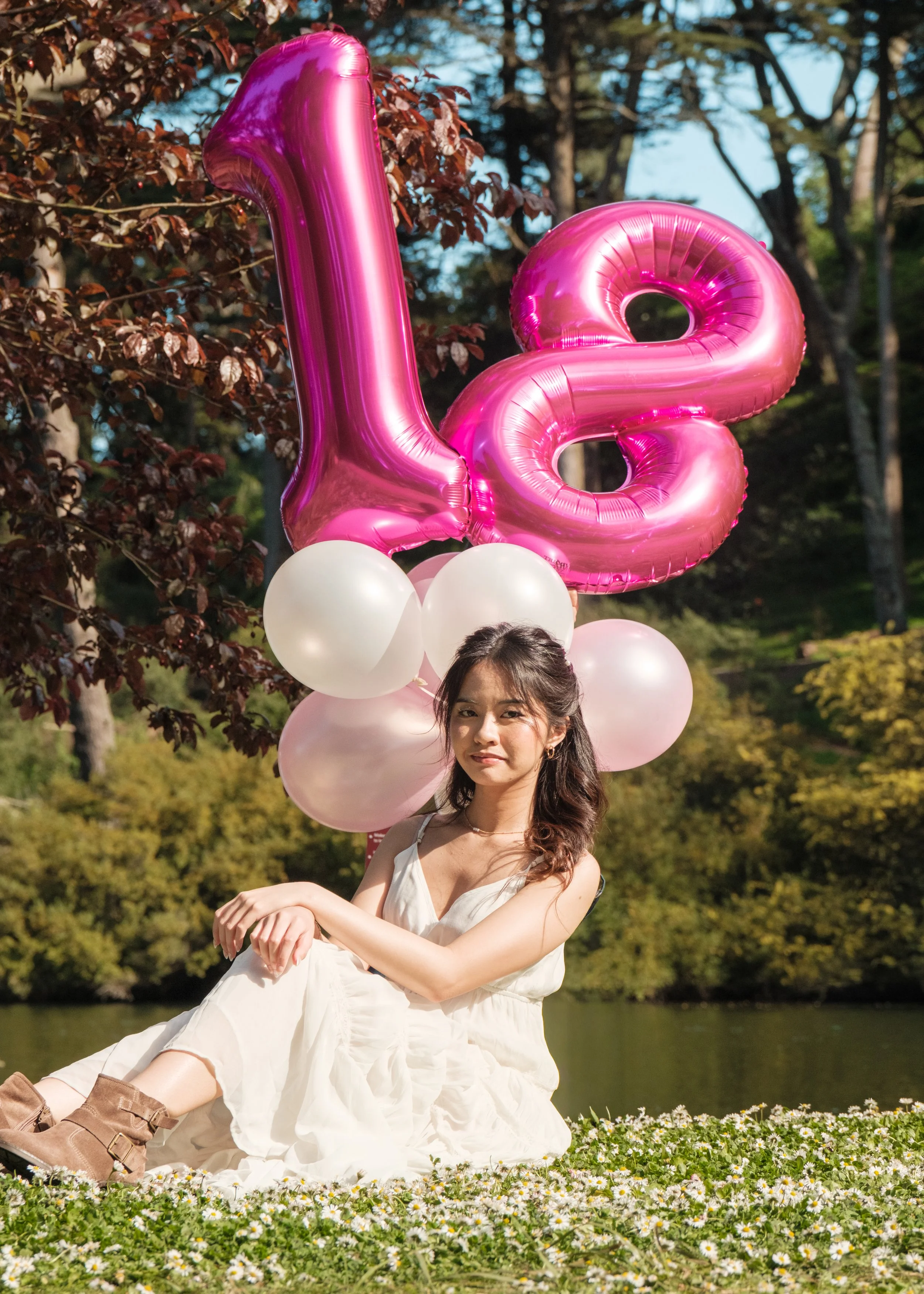 A young woman sitting outdoors on grass with white flowers, holding pink and white balloons that spell out the number 18, celebrating her 18th birthday.