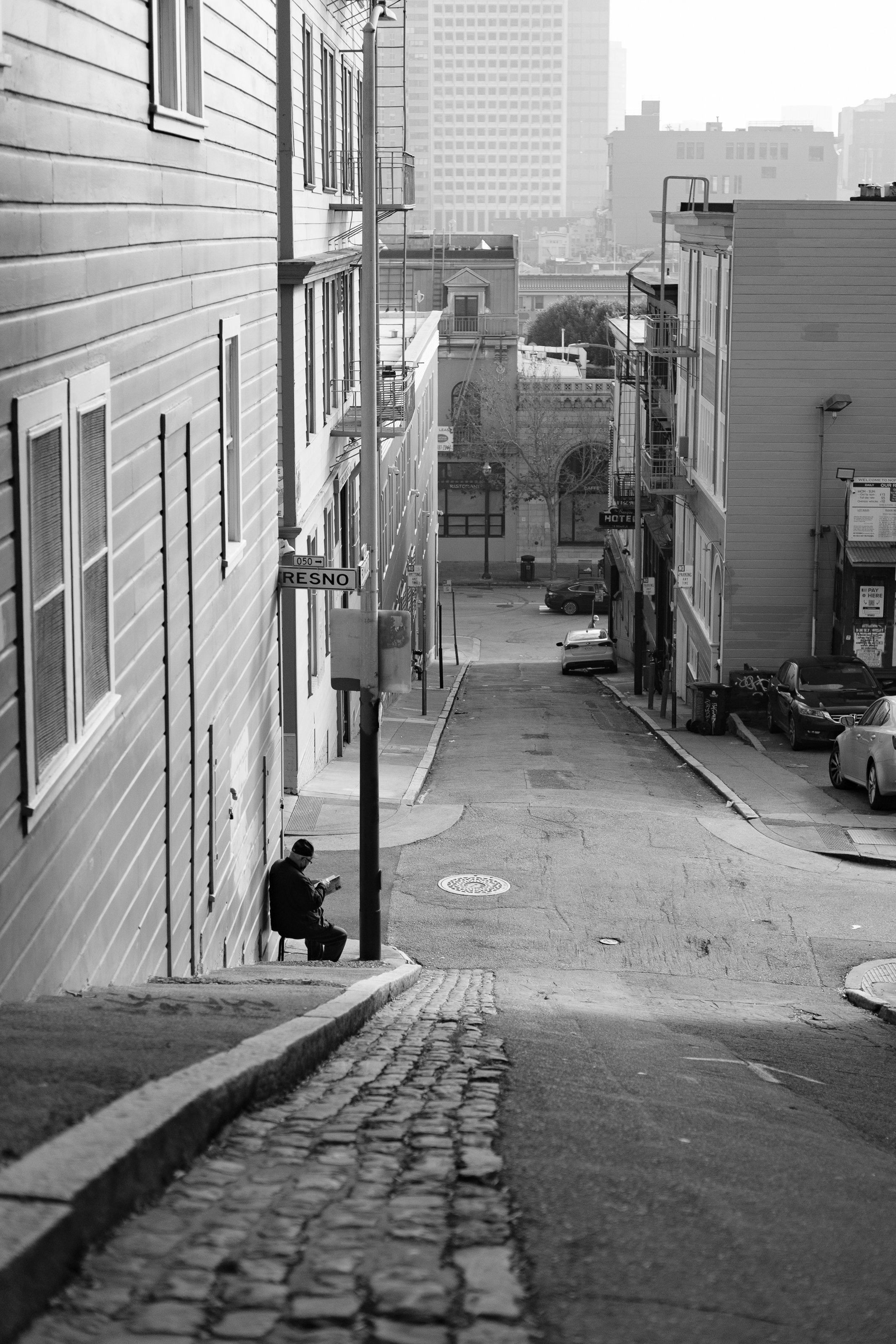 A black and white photo of an urban street scene with a man sitting on a bench and reading a book, overlooking a steep street with parked cars and tall buildings in the background.