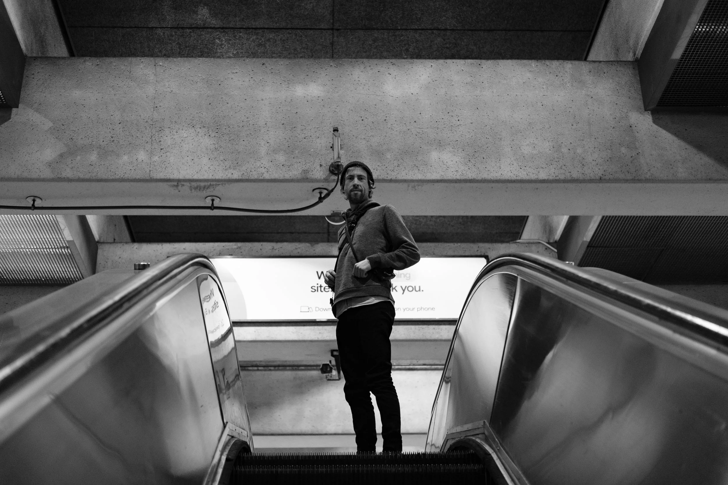 A man standing on an escalator in an indoor public space, looking at the camera, with concrete walls and a bright sign behind him.