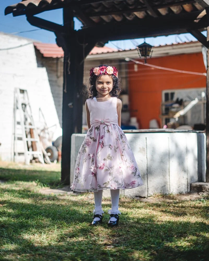 A young girl in a pink floral dress with a bow and a flower crown, standing outdoors on grass, smiling at the camera under a wooden structure.