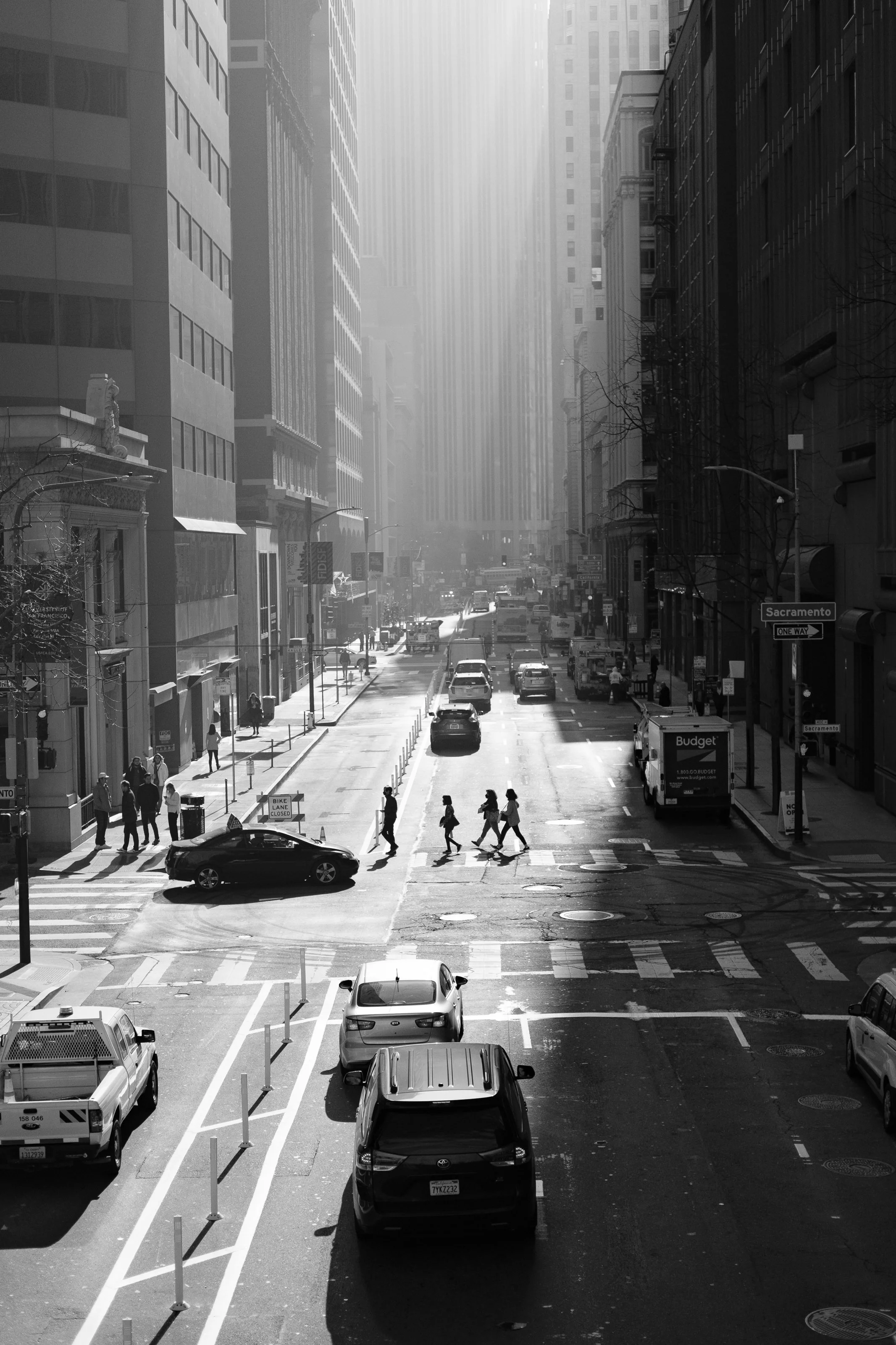A city street scene in black and white showing cars parked and moving on the road, pedestrians crossing at a crosswalk, tall buildings on both sides, with sunlight streaming down from above.