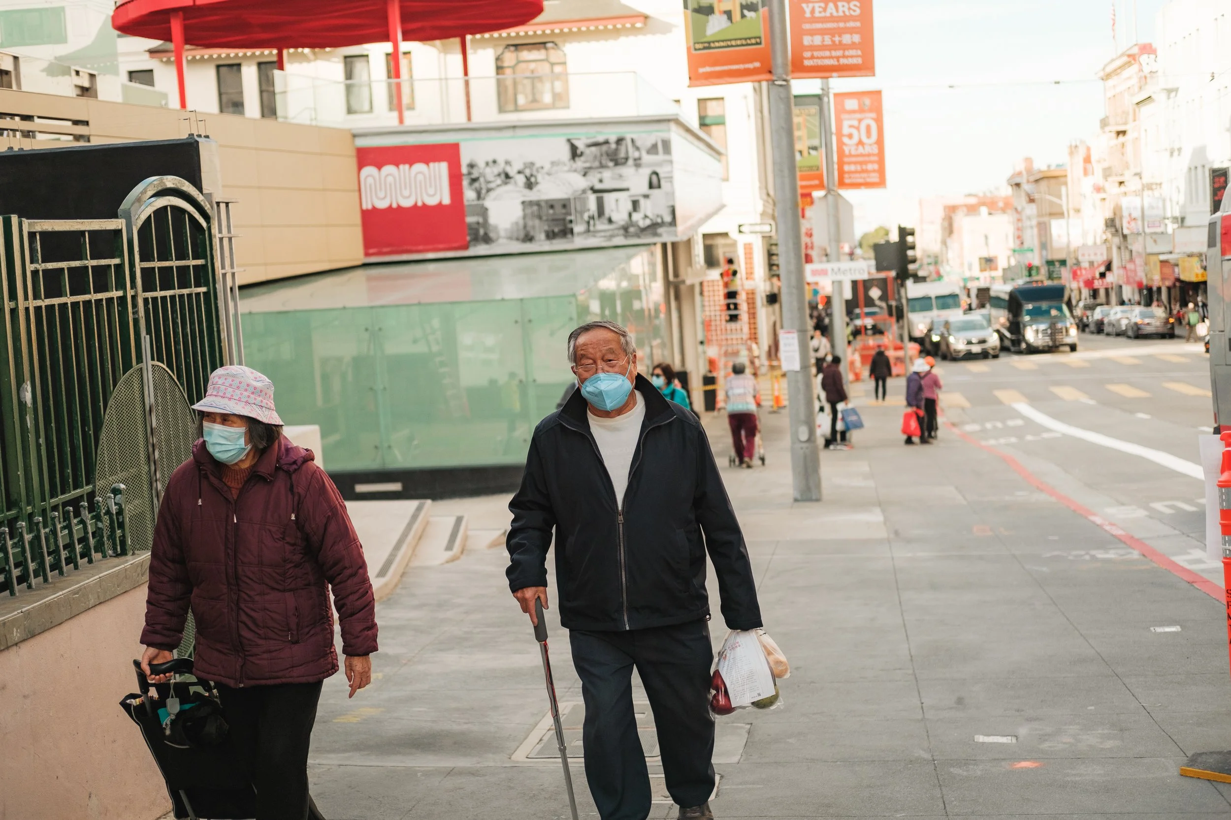 Two elderly individuals wearing masks walk on a busy city sidewalk. One woman with a hat and maroon jacket uses a walker, and a man with a cane carries groceries. In the background, there is street traffic, pedestrians, and buildings with promotional