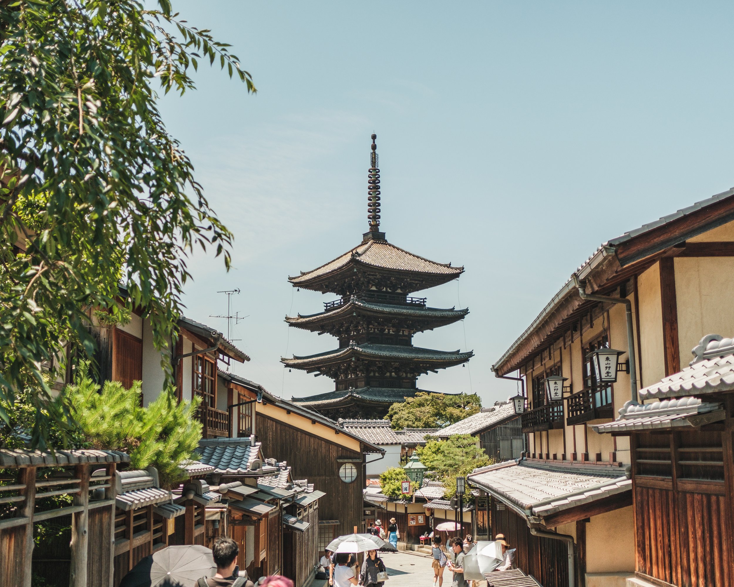 Traditional Japanese street with wooden buildings and a pagoda in the background, sunny day with people holding umbrellas.