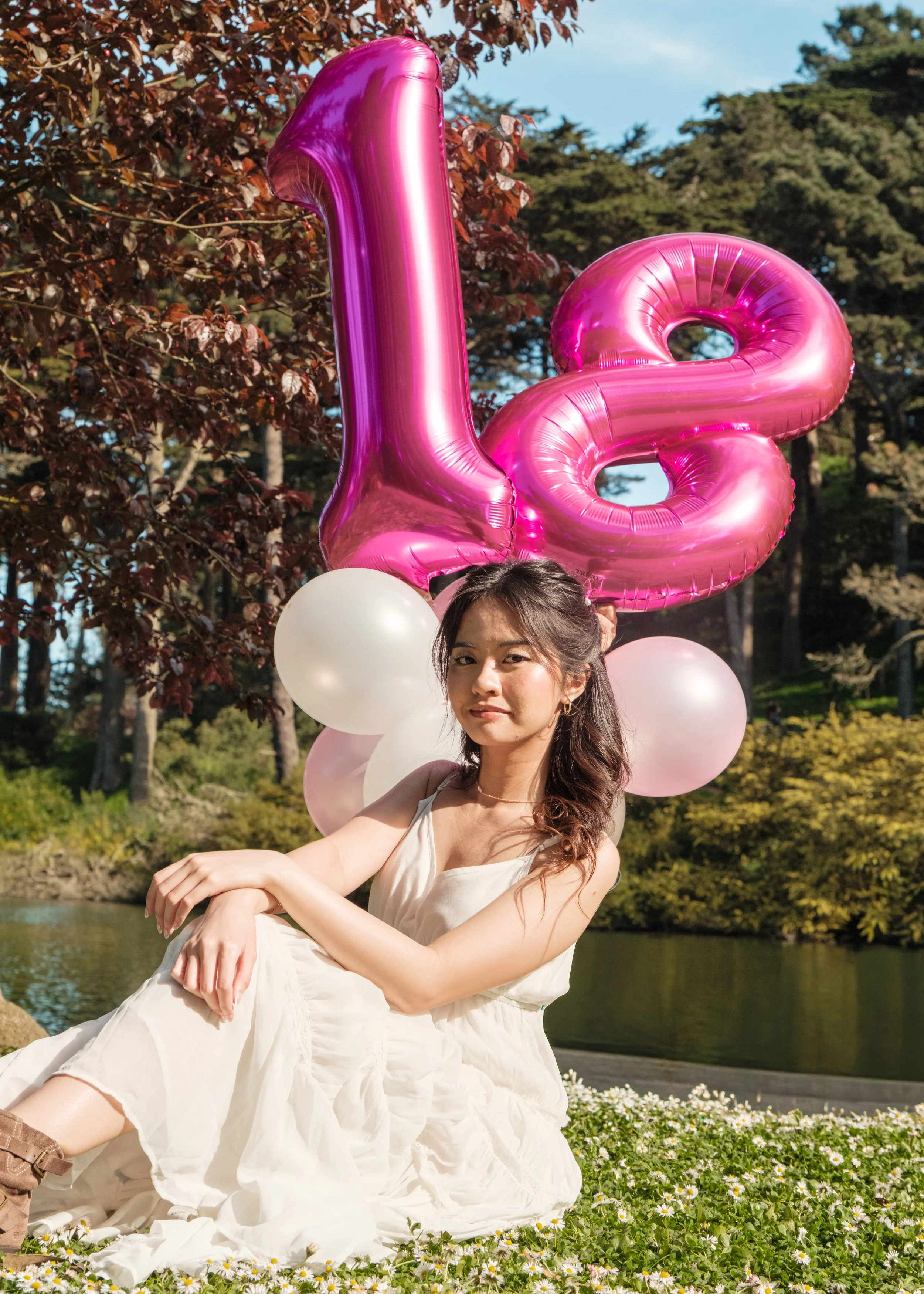 A young woman wearing a white dress sitting on grass with small white flowers, surrounded by balloons, including pink 'LOVE' letter balloons, near a pond with trees in the background on a sunny day.