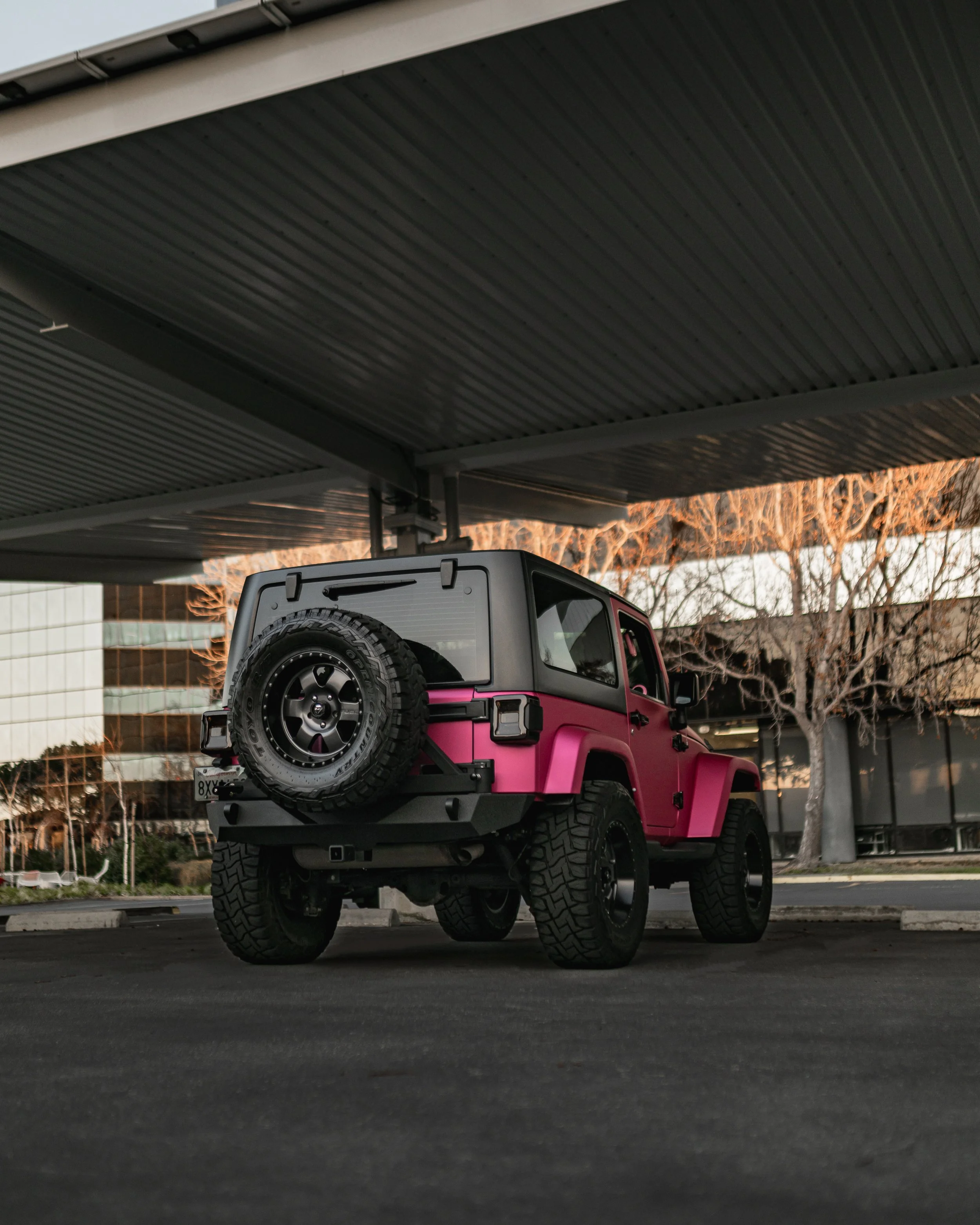 A pink off-road vehicle with a spare tire mounted on the rear, parked under a parking structure with an urban cityscape in the background.