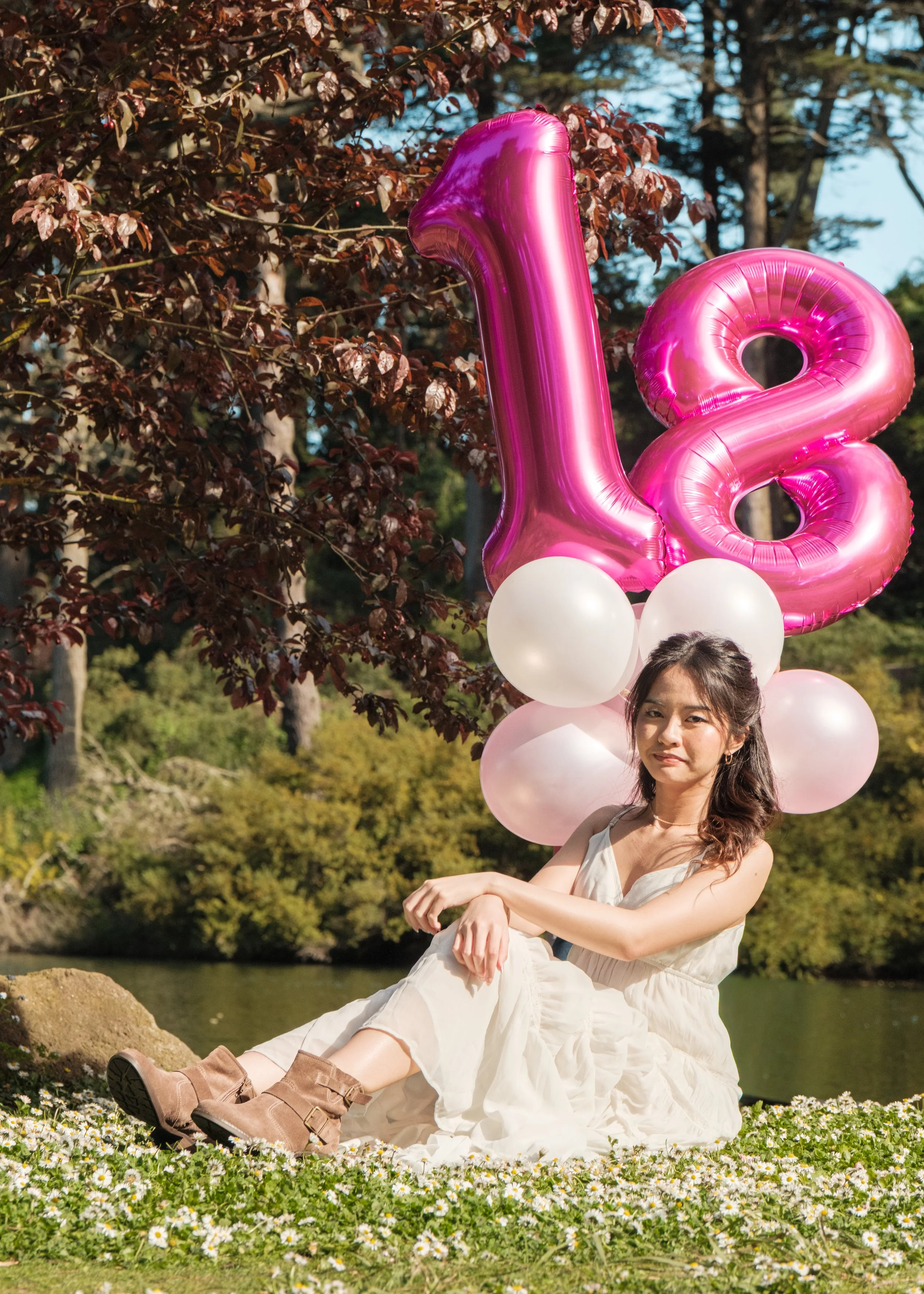 A young woman sits on a grassy field with white flowers, holding her knees, next to pink and white balloons. There are pink balloon numbers '1' and '8'. She is outdoors with trees in the background and the day appears sunny.