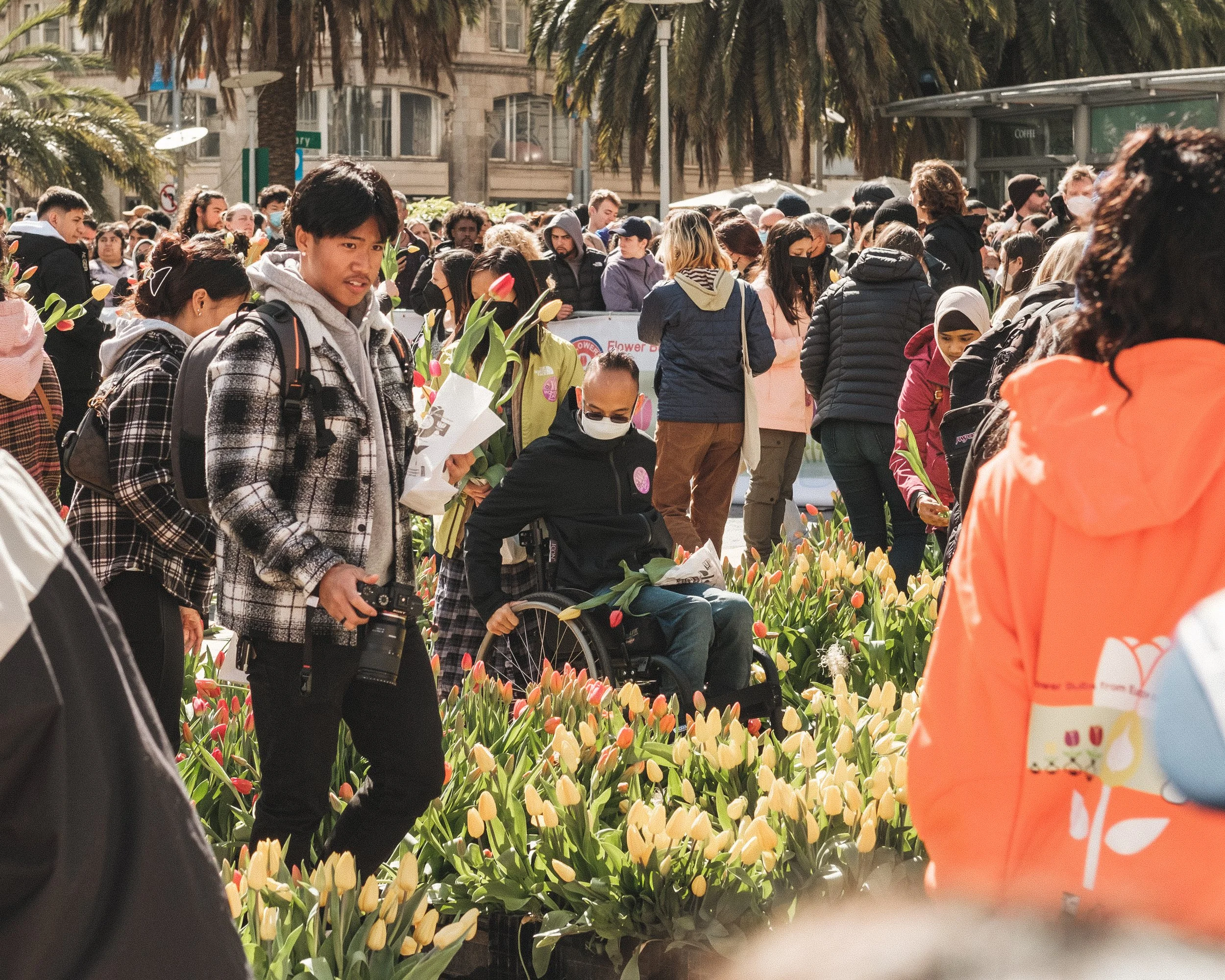 A crowd of people, some holding tulips, gathered at an outdoor flower market or event with palm trees and buildings in the background on a sunny day.