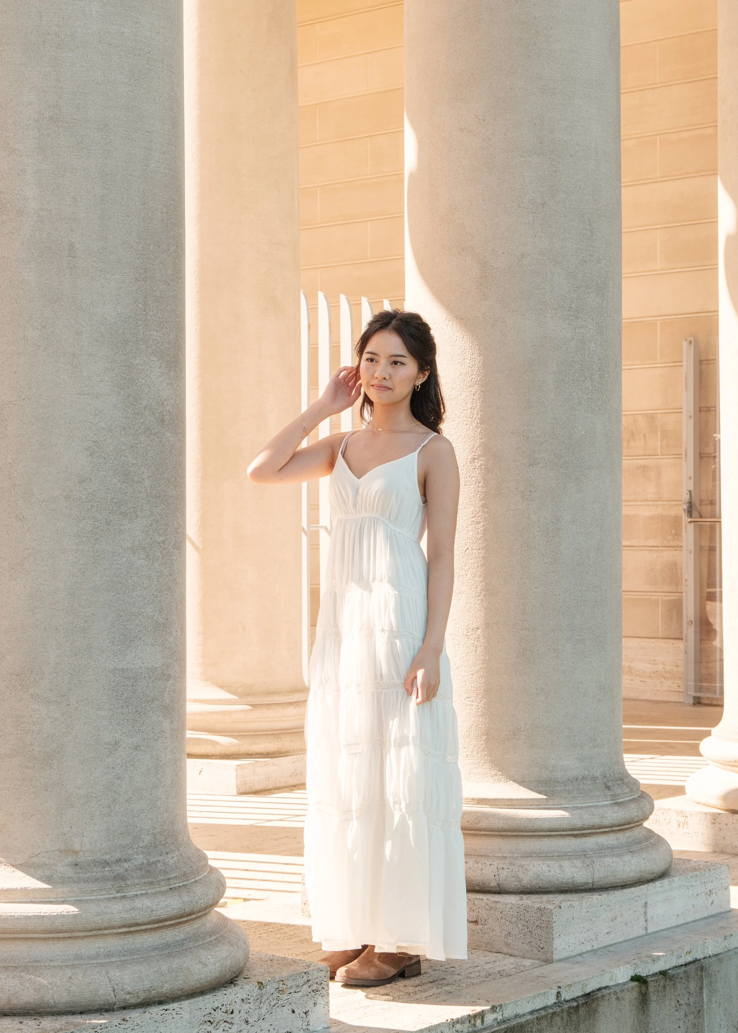 A young woman in a white summer dress standing among large classical columns outside during the daytime.