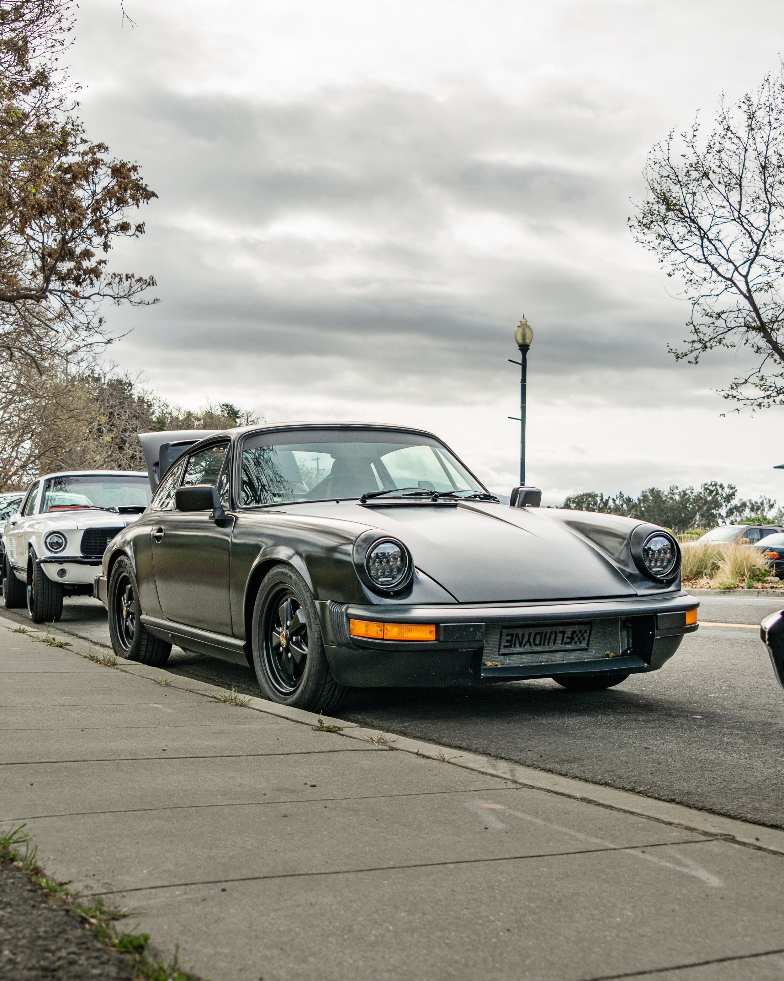 A black vintage Porsche 911 sports car parked on a street, with other cars behind it and a cloudy sky above.