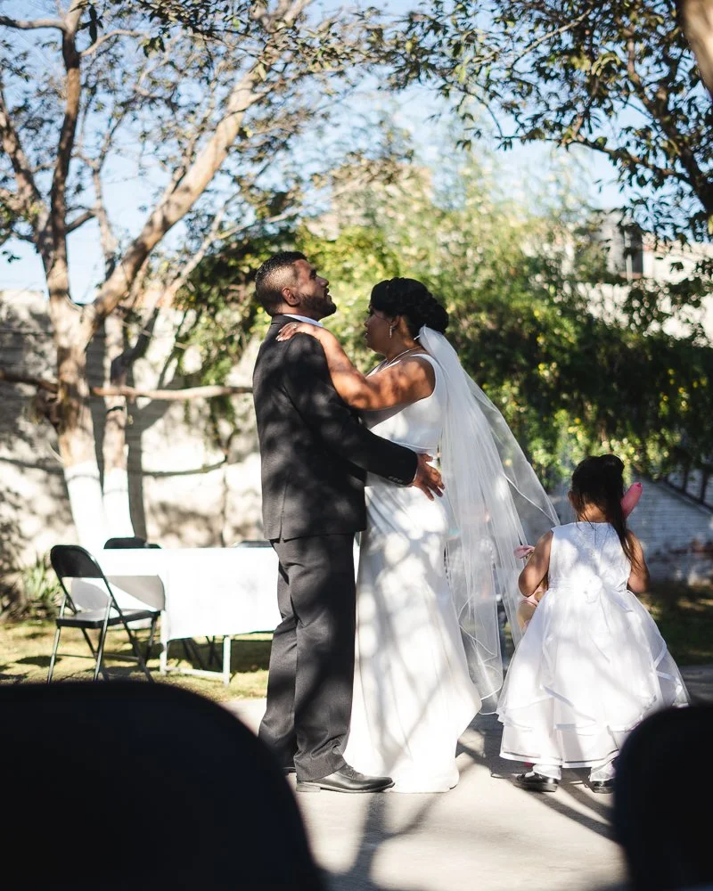 A couple in wedding attire embracing outdoors, a young girl in a white dress standing nearby, with trees and a table in the background.