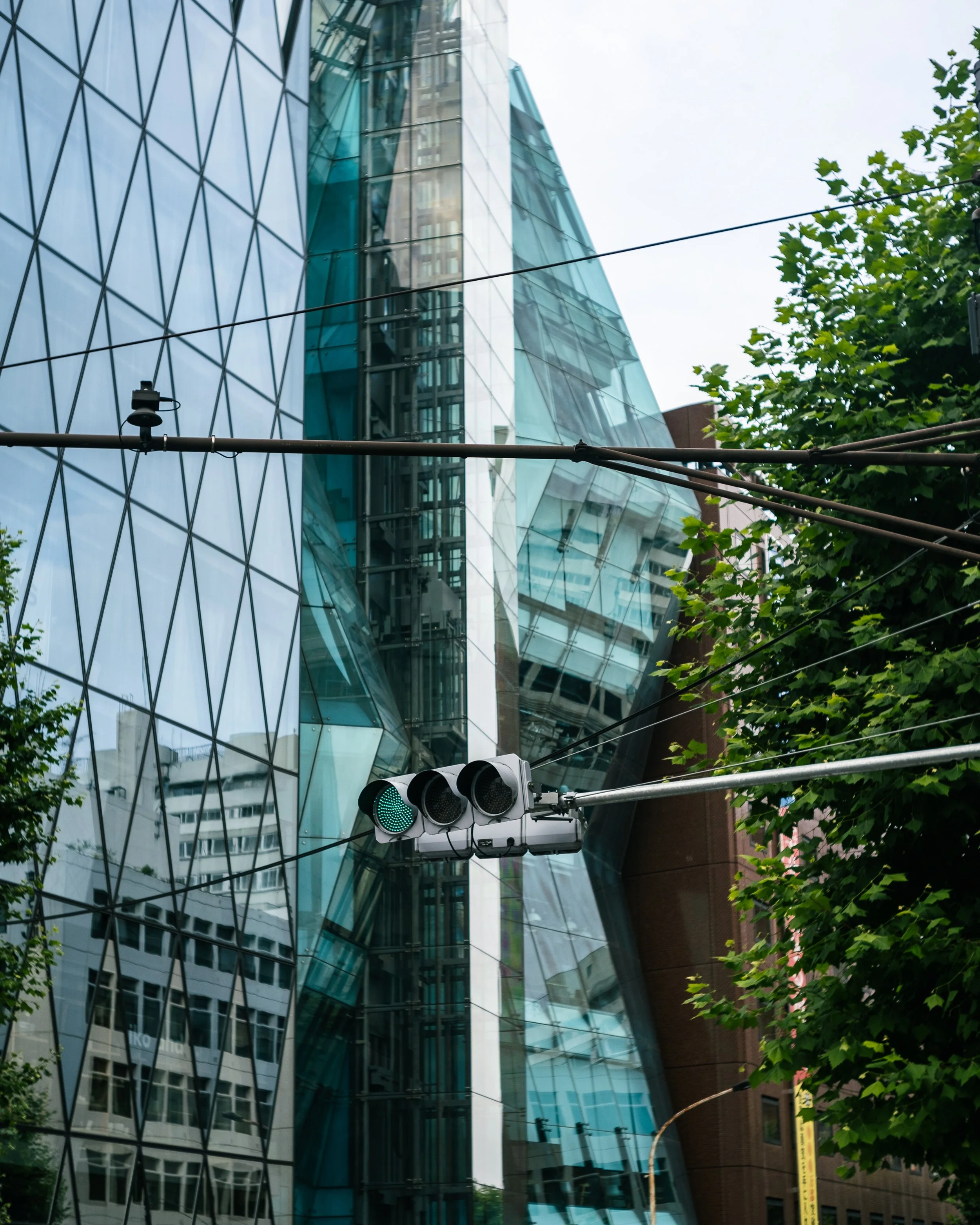 A city street scene with a modern glass building, a traffic light showing green, and trees partially blocking the view.