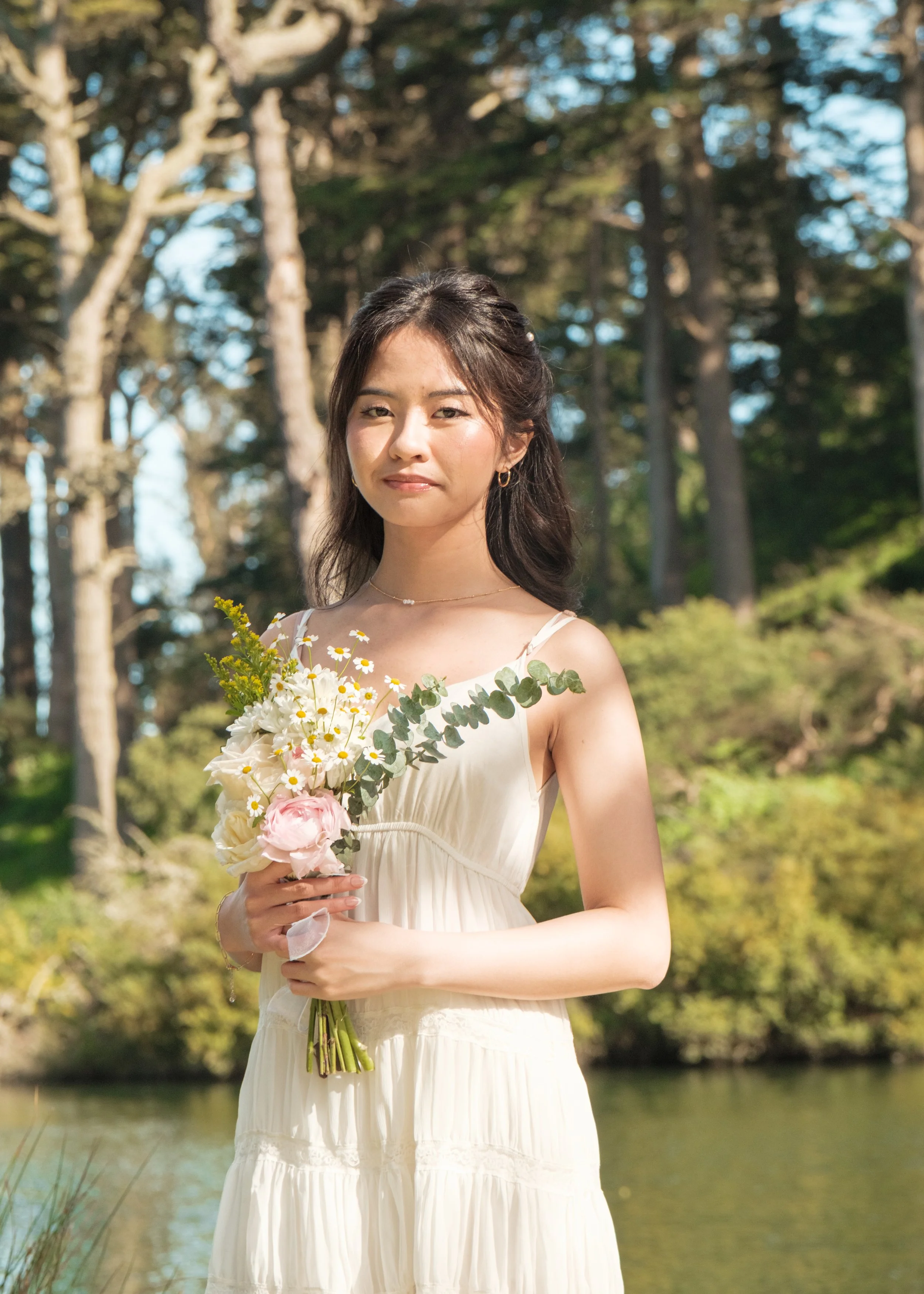 A young woman in a white dress outdoors holding a bouquet of flowers, with trees and a body of water in the background.