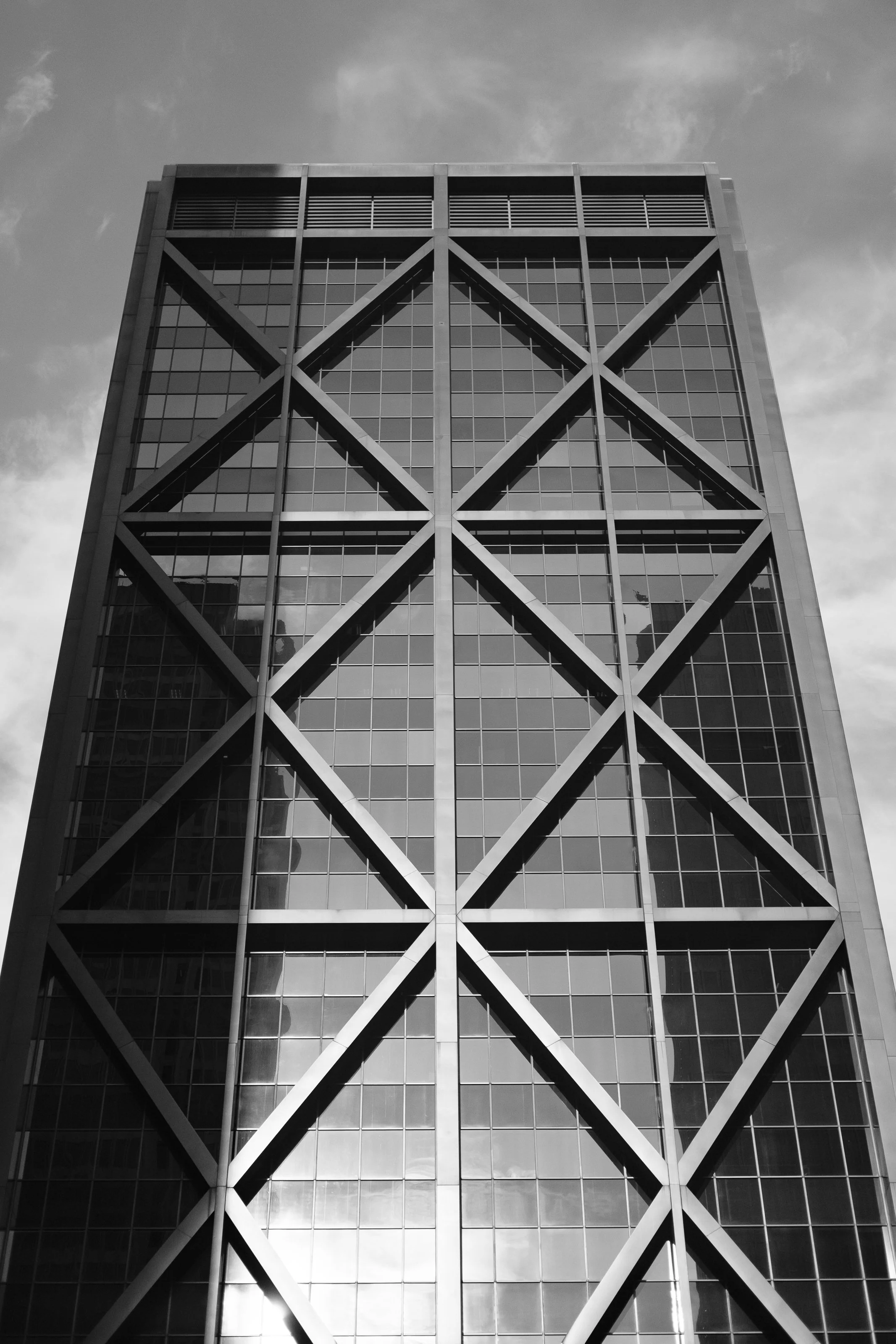 Black and white photograph of a tall modern glass office building with prominent crossbeam structural design on its facade, viewed from ground level looking up.