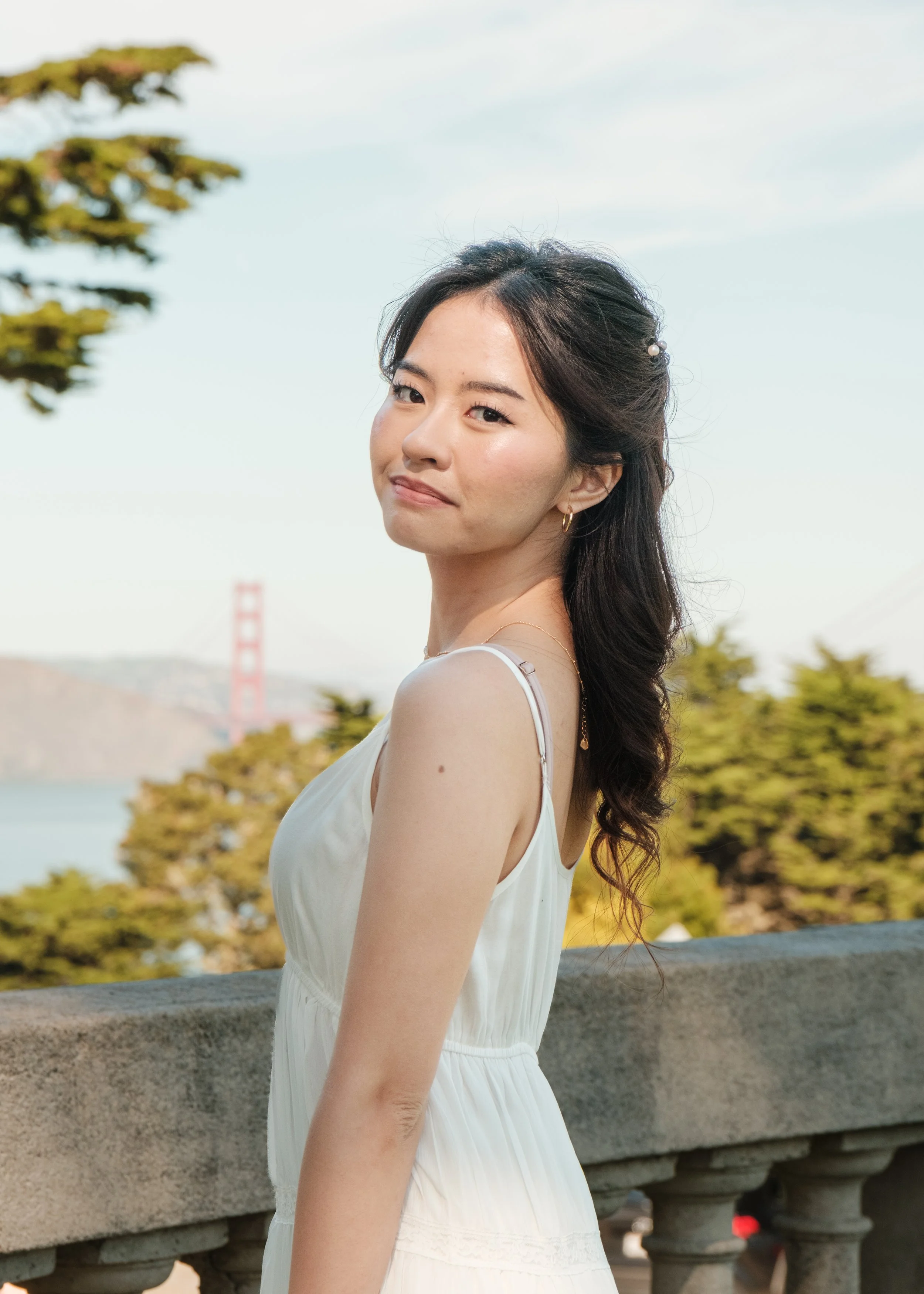 A young woman with long dark hair, wearing a sleeveless white dress, standing outdoors near a stone railing with trees in the background and a bridge visible in the distance.