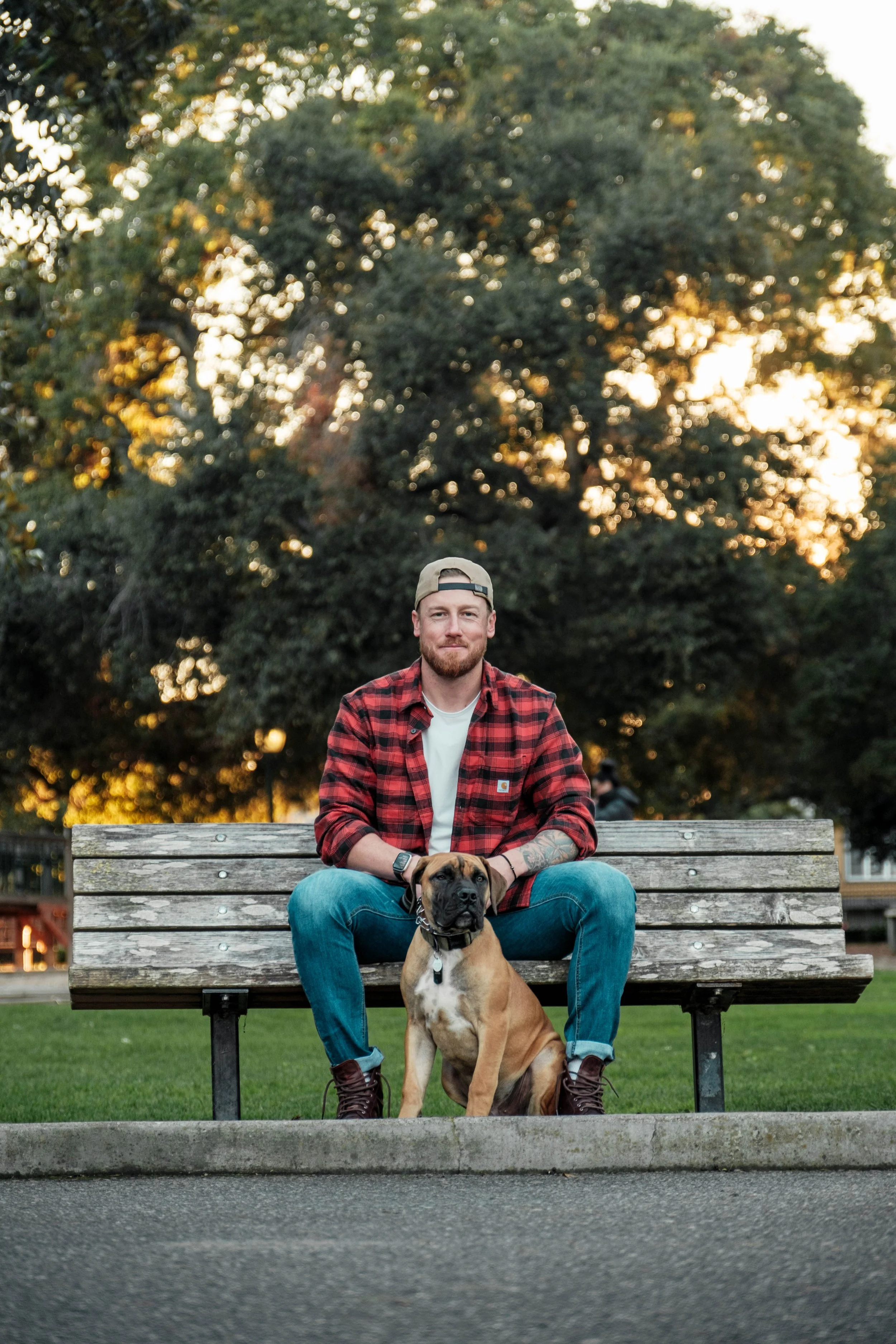 A man with a beard and tattoos sitting on a park bench with a brown dog in front of him, with trees in the background during sunset.