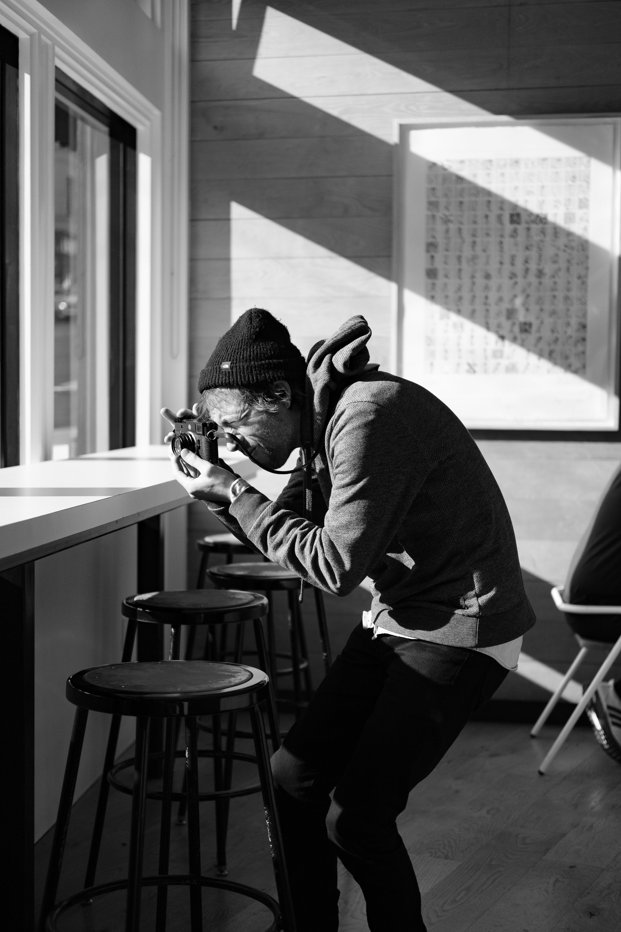 A man in a beanie and hoodie taking a photograph inside a room with wooden walls, sunlight casting shadows, and empty stools along a counter.