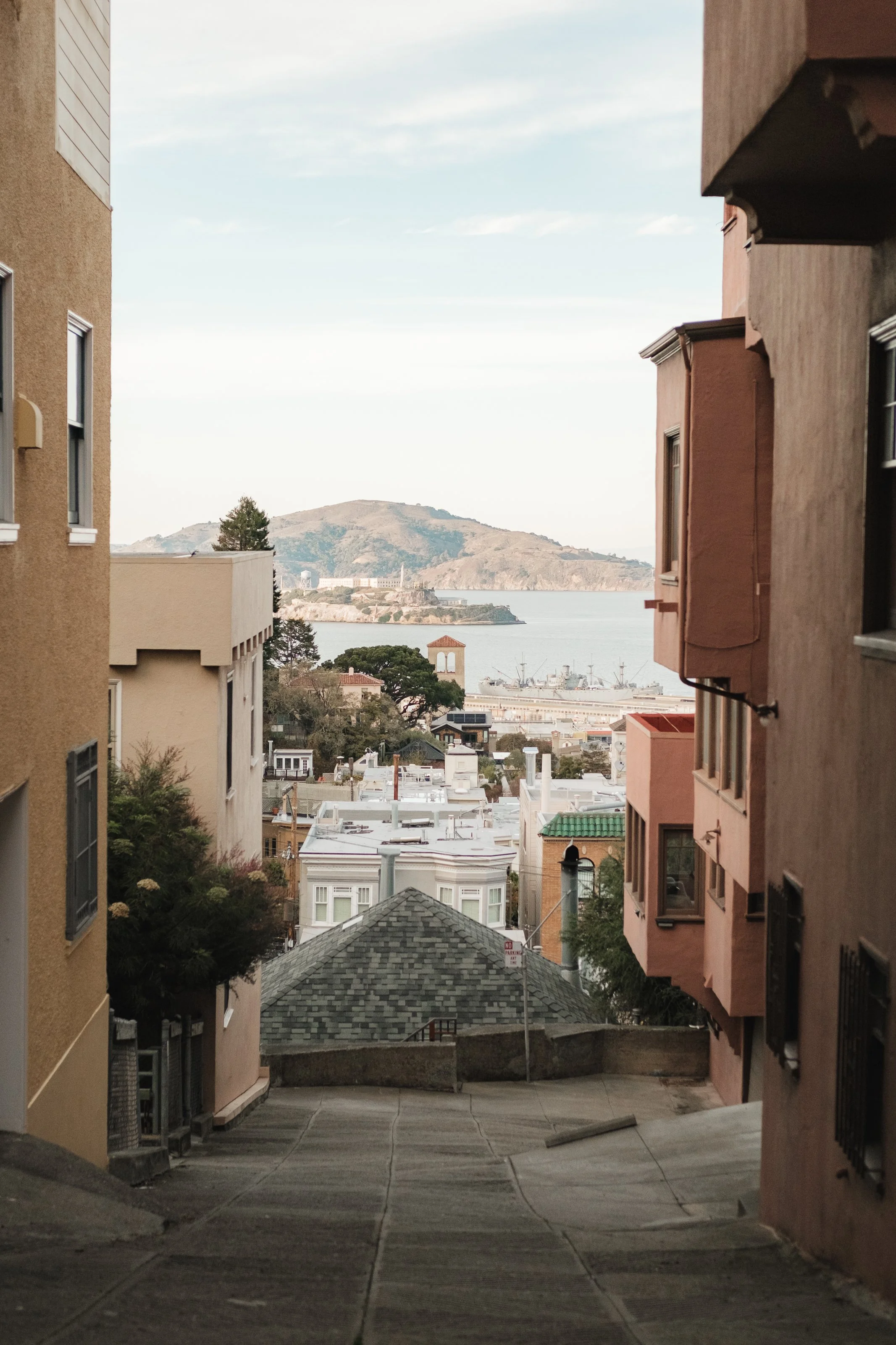 View down a steep street between colorful apartment buildings towards a waterfront with ships and hills in the distance.