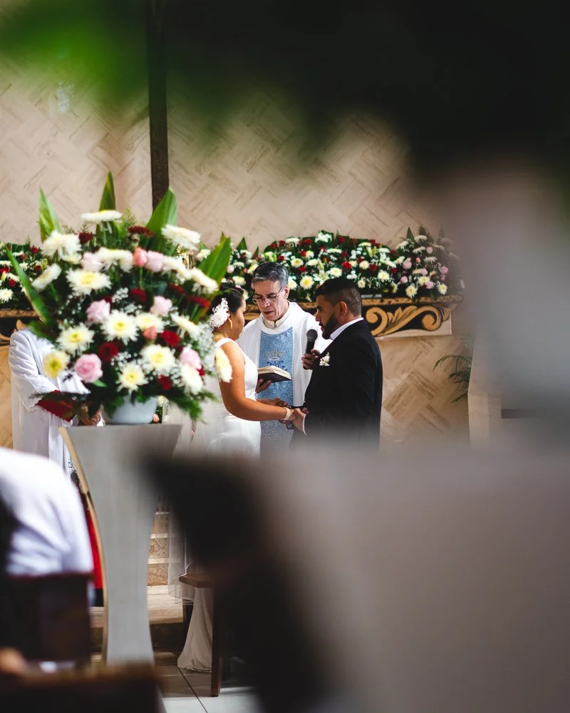 A couple getting married in a church, standing before a priest, holding hands, with a large floral arrangement nearby and more flowers in the background.