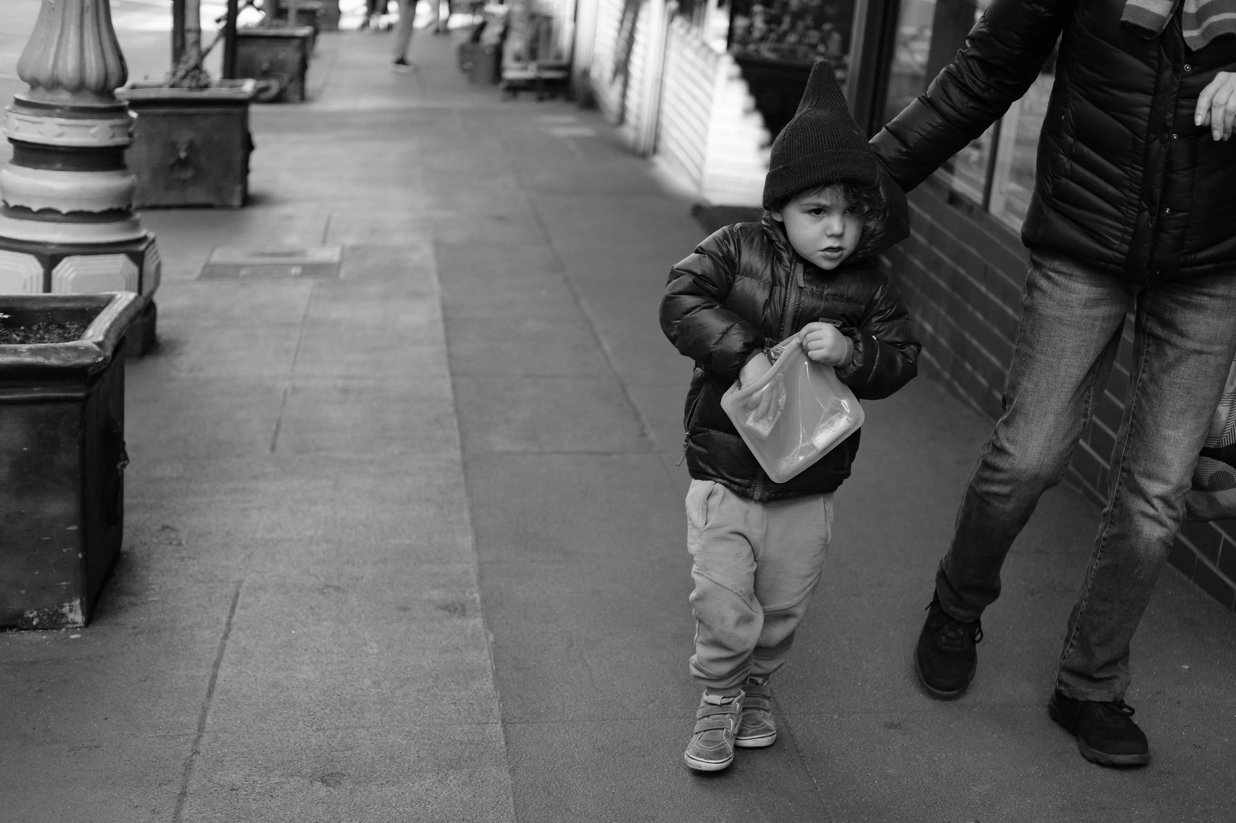 A young boy wearing a black jacket, beige pants, and a beanie hat walking on a sidewalk holding a plastic container, with an adult partially visible next to him. They are in an urban area with storefronts and planters lining the sidewalk.