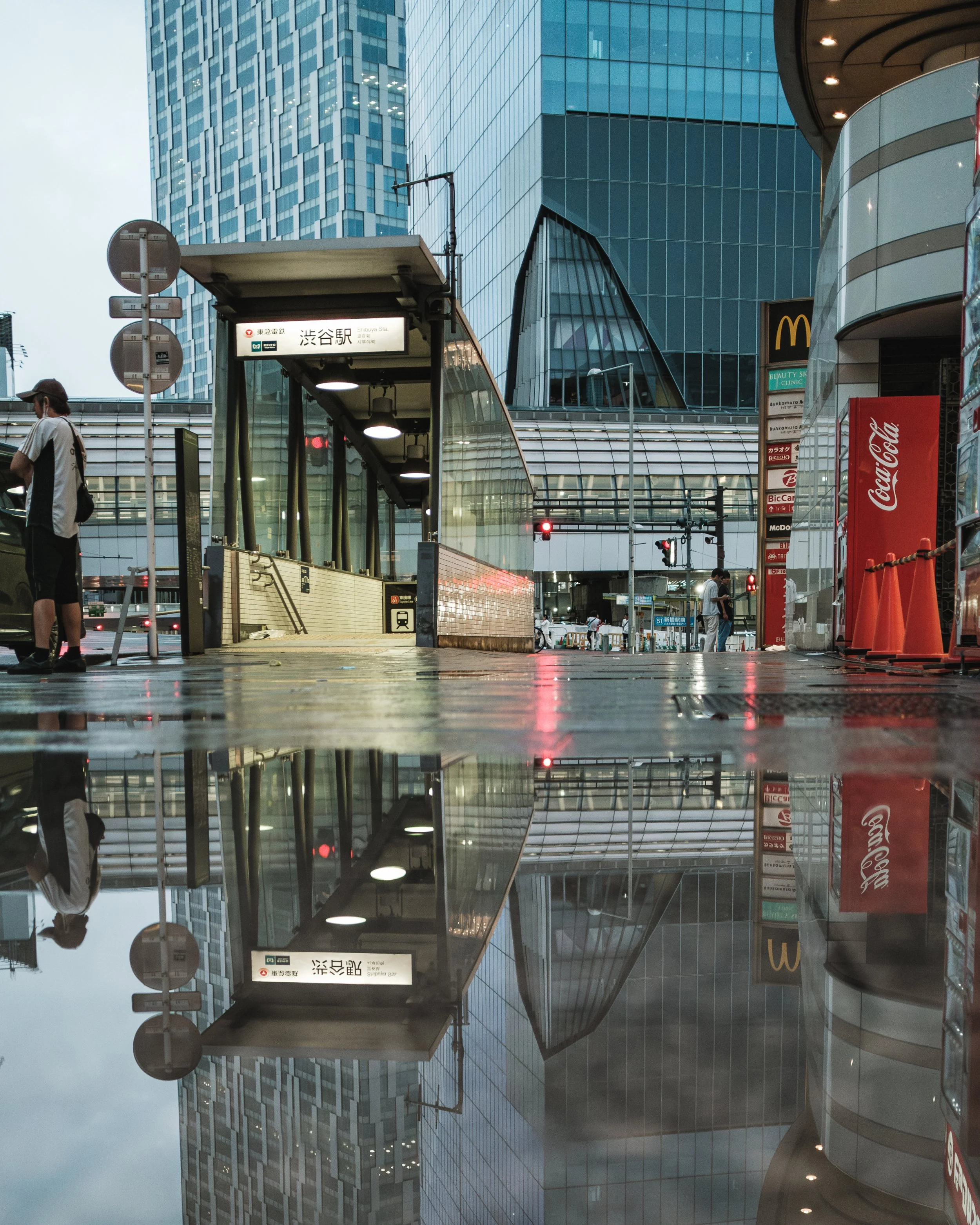 City street scene at Shibuya Station in Tokyo, Japan with modern glass buildings, an entrance to the subway, people walking, and reflections on wet pavement