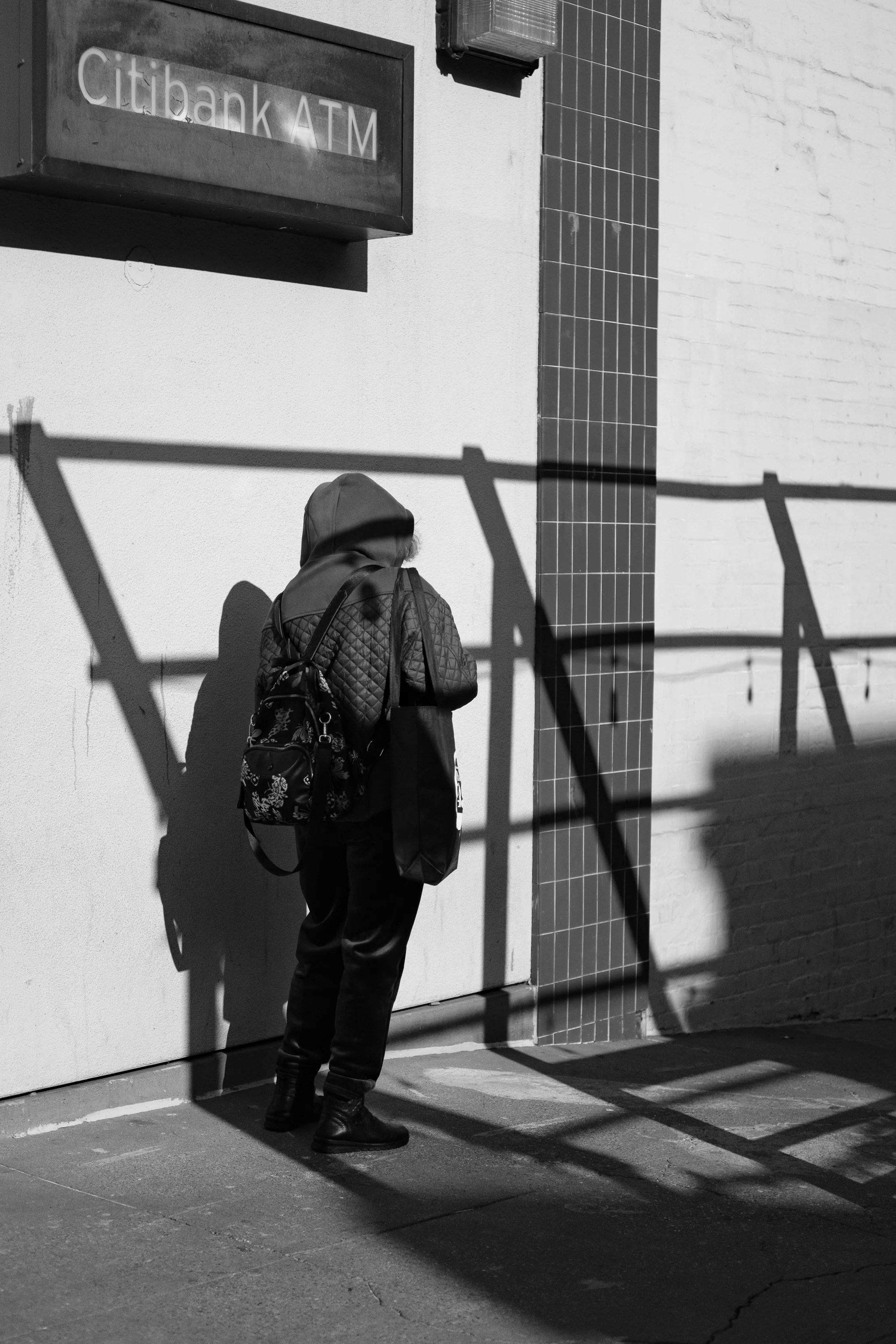 A person in a hoodie and dark clothing stands outside a building with a sign that reads 'citibank ATM'. The person has a backpack and is casting a shadow on a white wall, with shadows of a metal railing crossing the scene.