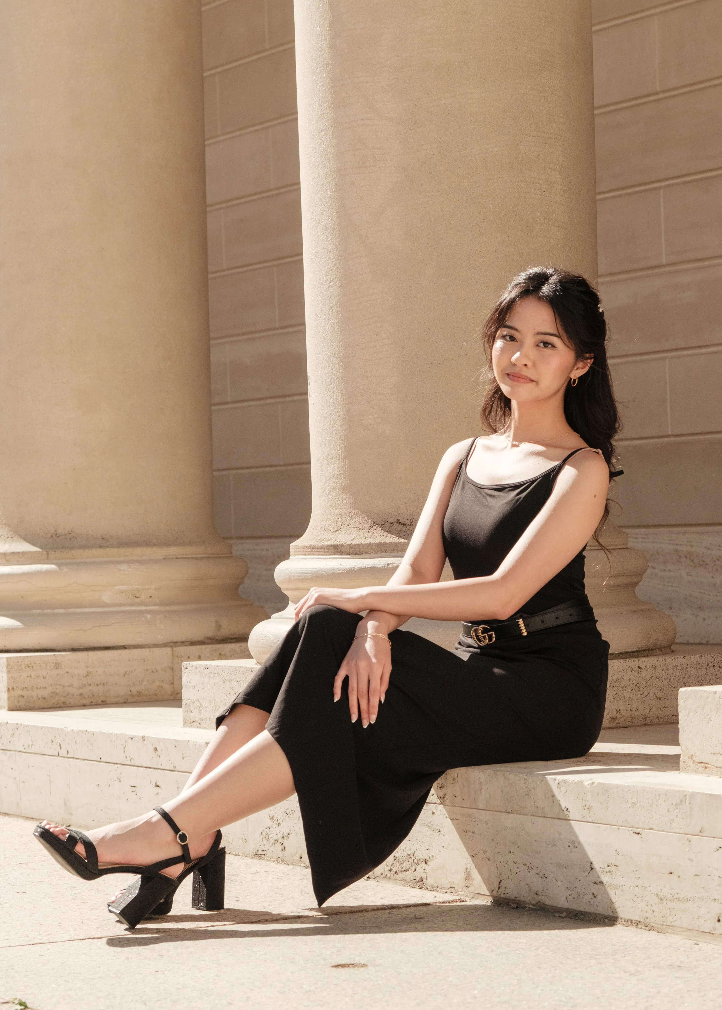 A woman with long dark hair sitting on stairs in front of large columns, wearing a black sleeveless dress and high-heeled sandals, posing outdoors in sunlight.