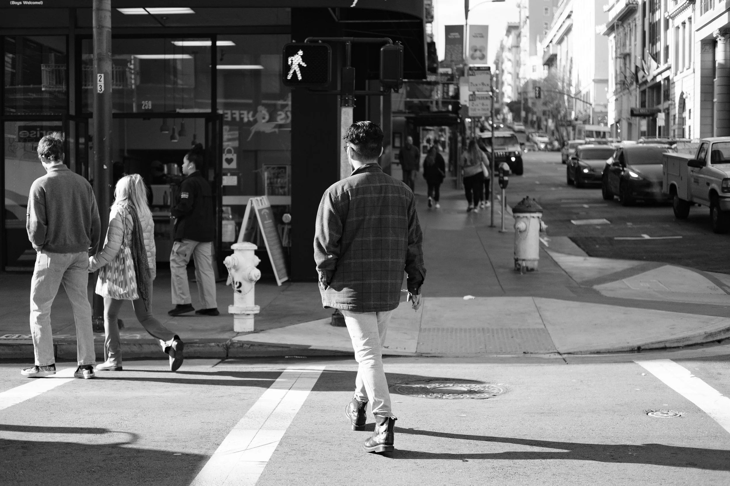 Street scene in an urban area with pedestrians crossing at a crosswalk, including a man in a jacket walking away, a boy and girl holding hands, and a woman in a jacket walking. Cars parked along the street and buildings line the sidewalk.