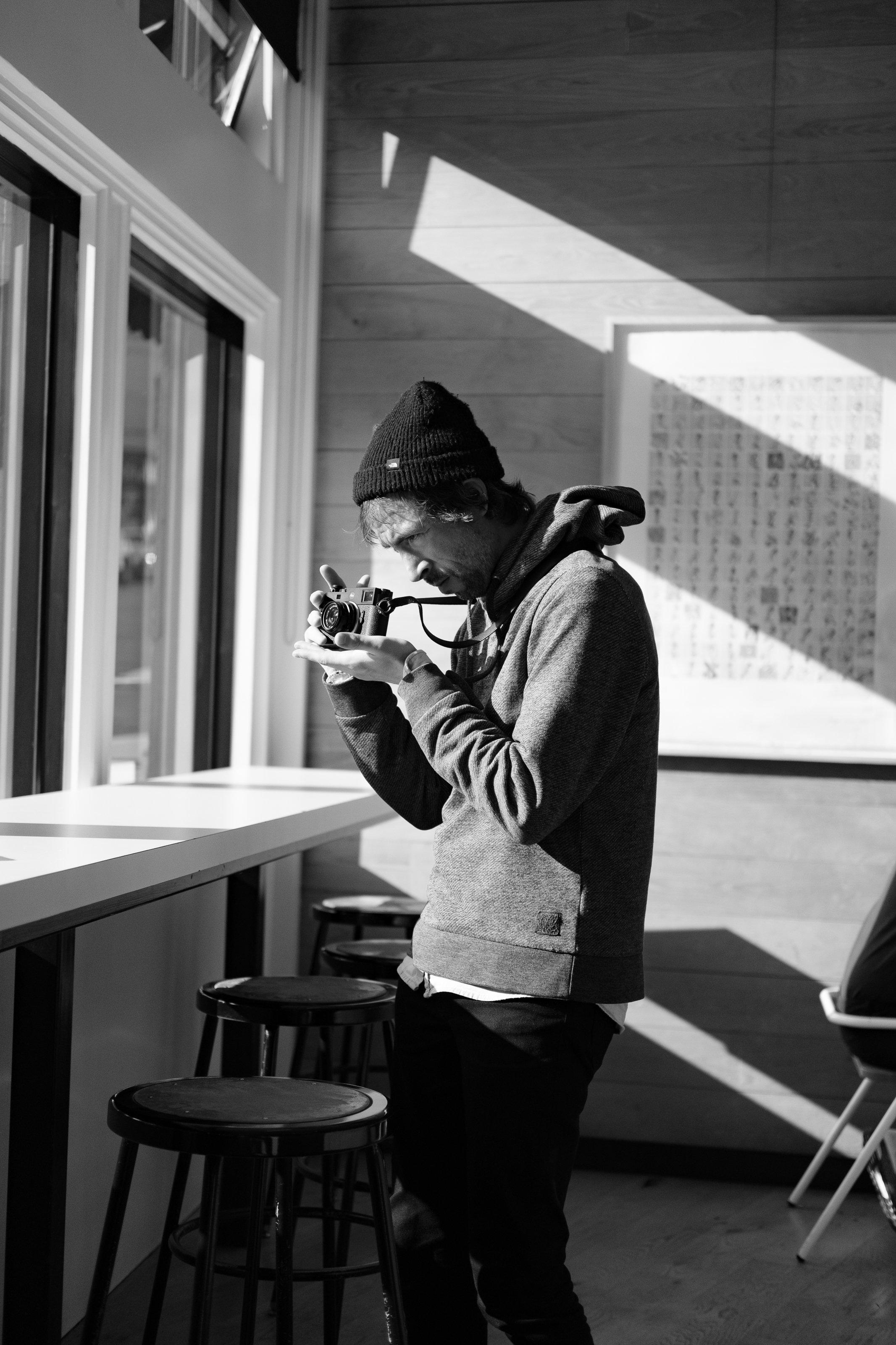 A man wearing a beanie and hoodie looking at a camera indoors with sunlight streaming through windows.