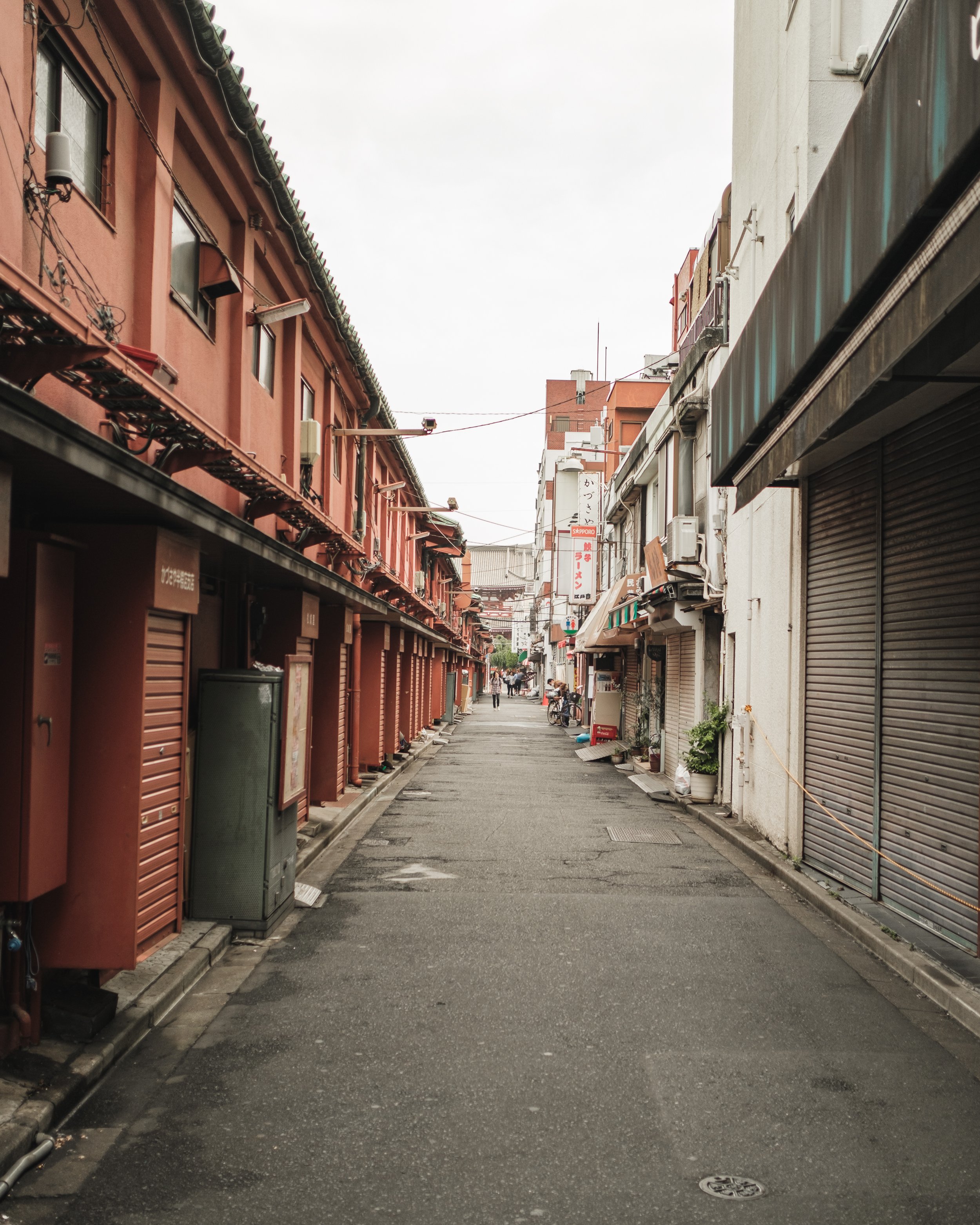 A quiet narrow street in an urban area with buildings on both sides, some with shuttered storefronts and awnings, and a few people in the distance.