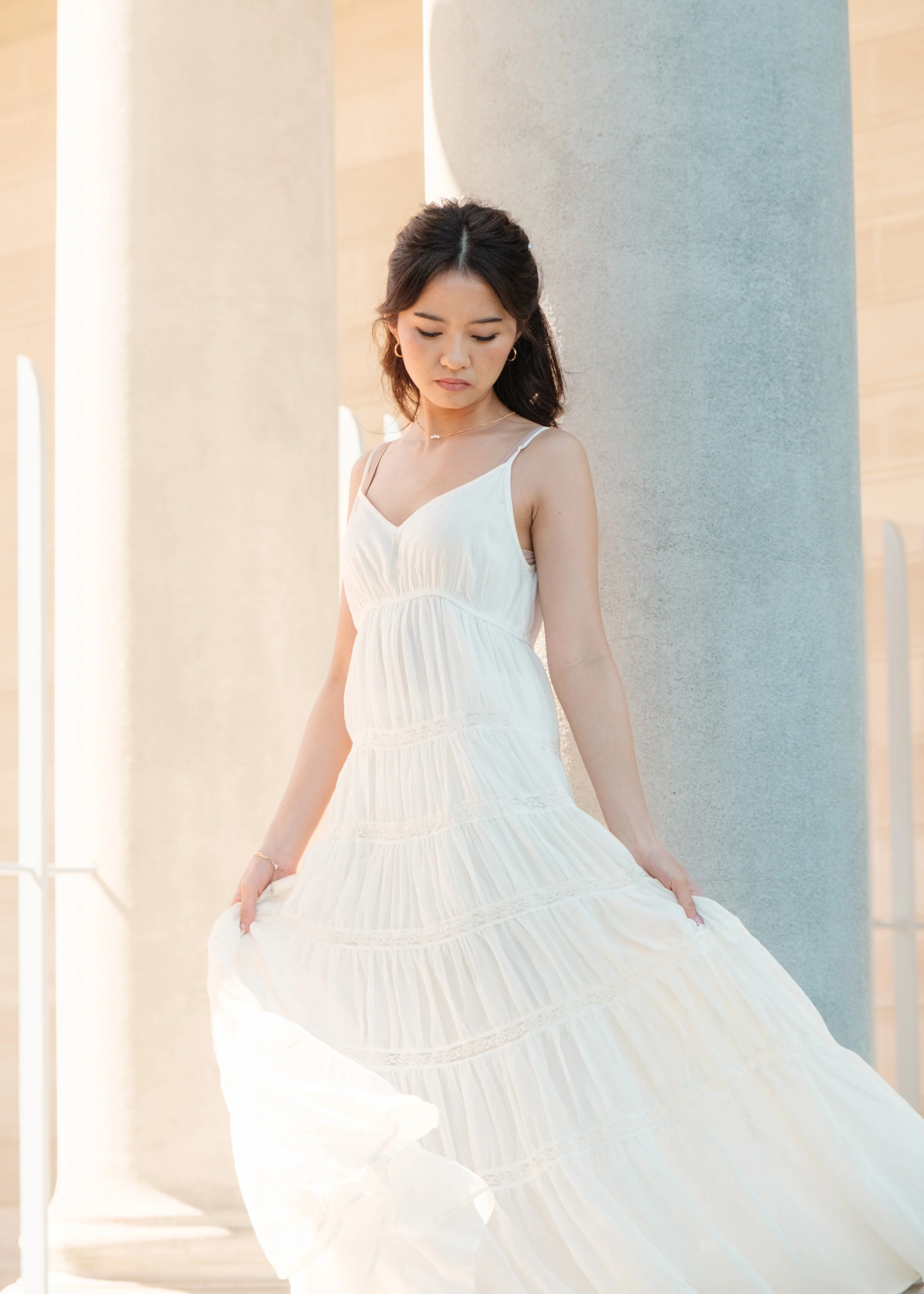 Young woman in a white, flowing, tiered maxi dress standing beside a large concrete pillar, looking down and holding the edge of her dress.