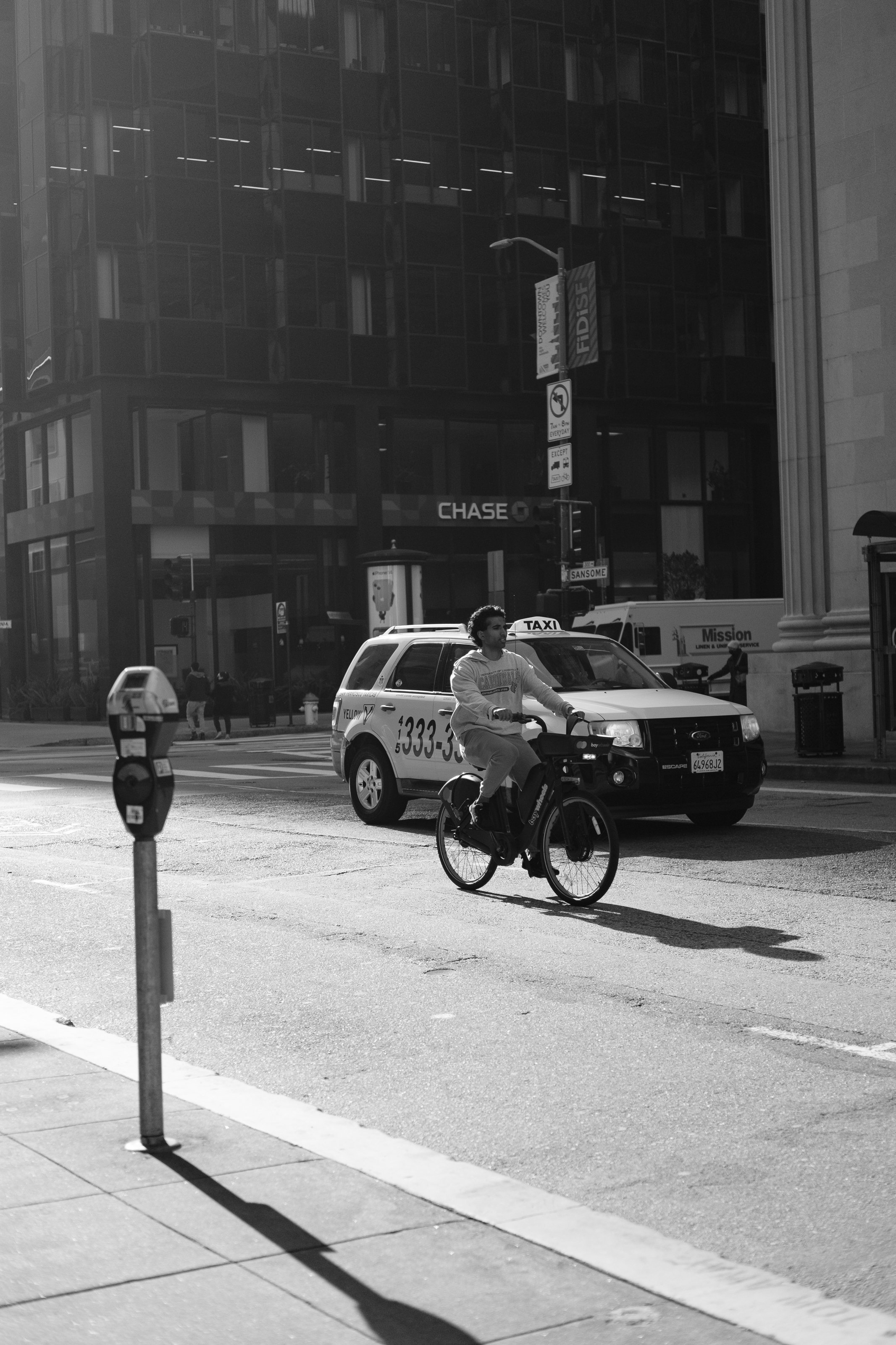 A person riding a bicycle on a city street near a parked taxi and a parking meter, with tall buildings in the background in black and white.