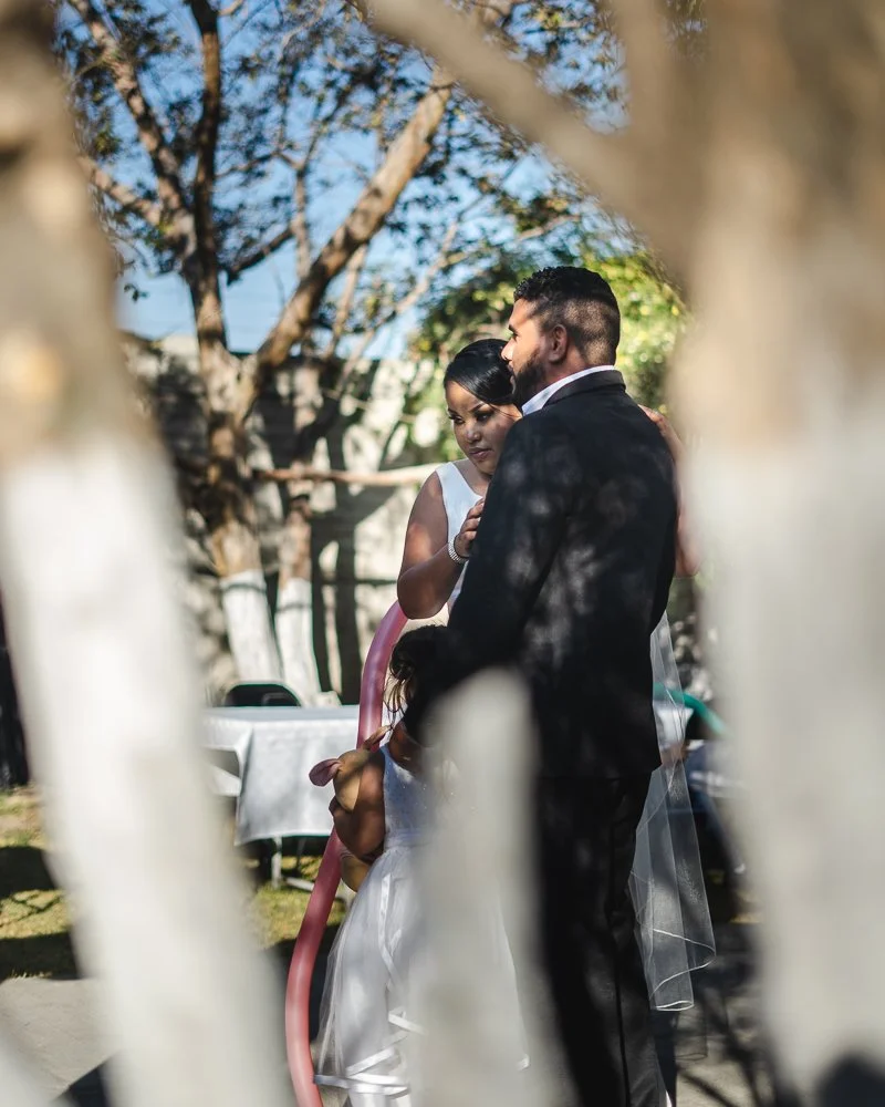 A couple dressed in wedding attire, with the woman in a white dress and the man in a suit, sharing a moment together outdoors, with a young girl in a white dress standing in front of them, partially obscured by a pink balloon arch.