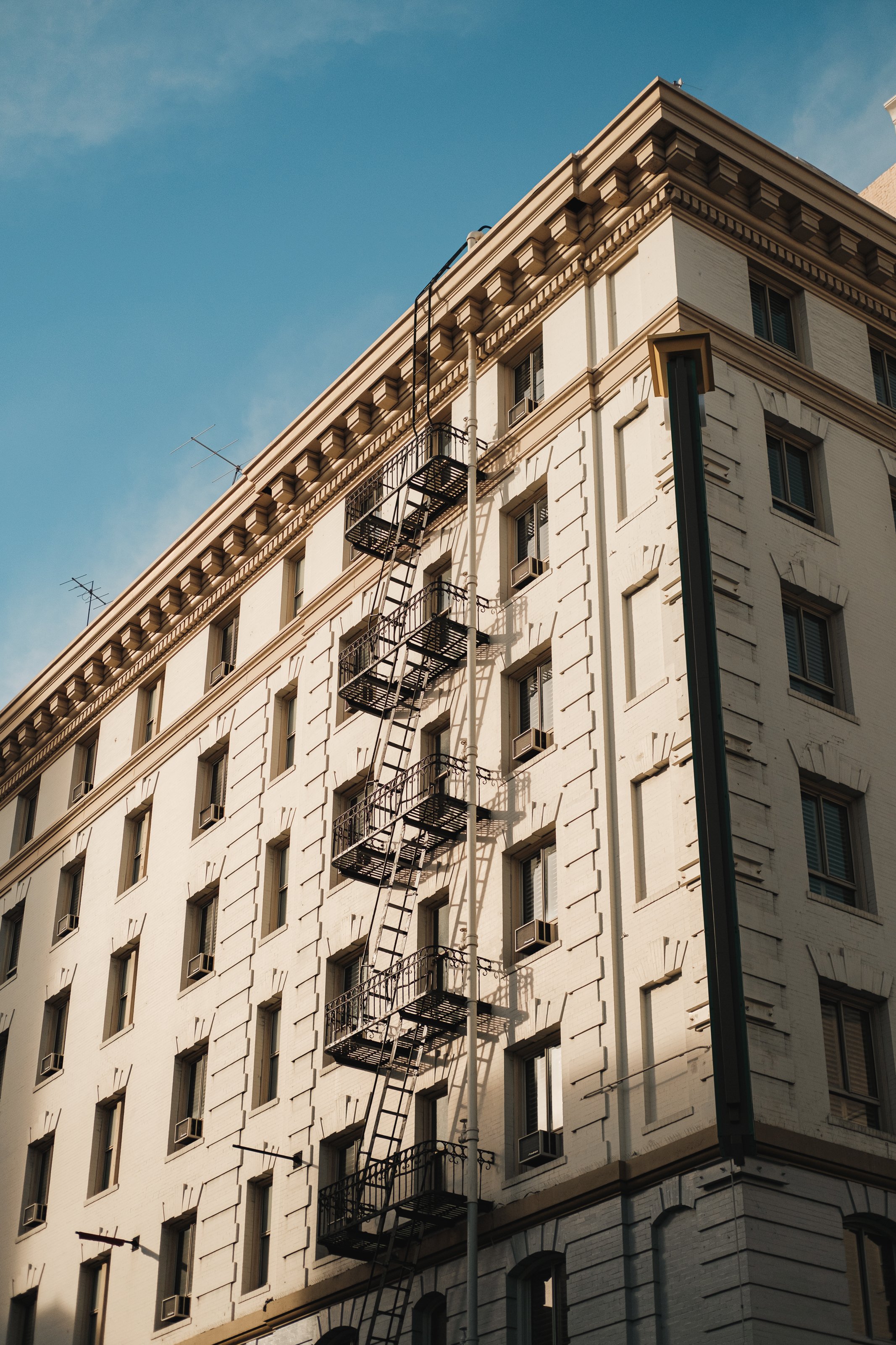 A tall white building with multiple windows and black fire escape stairs on the side, set against a blue sky.