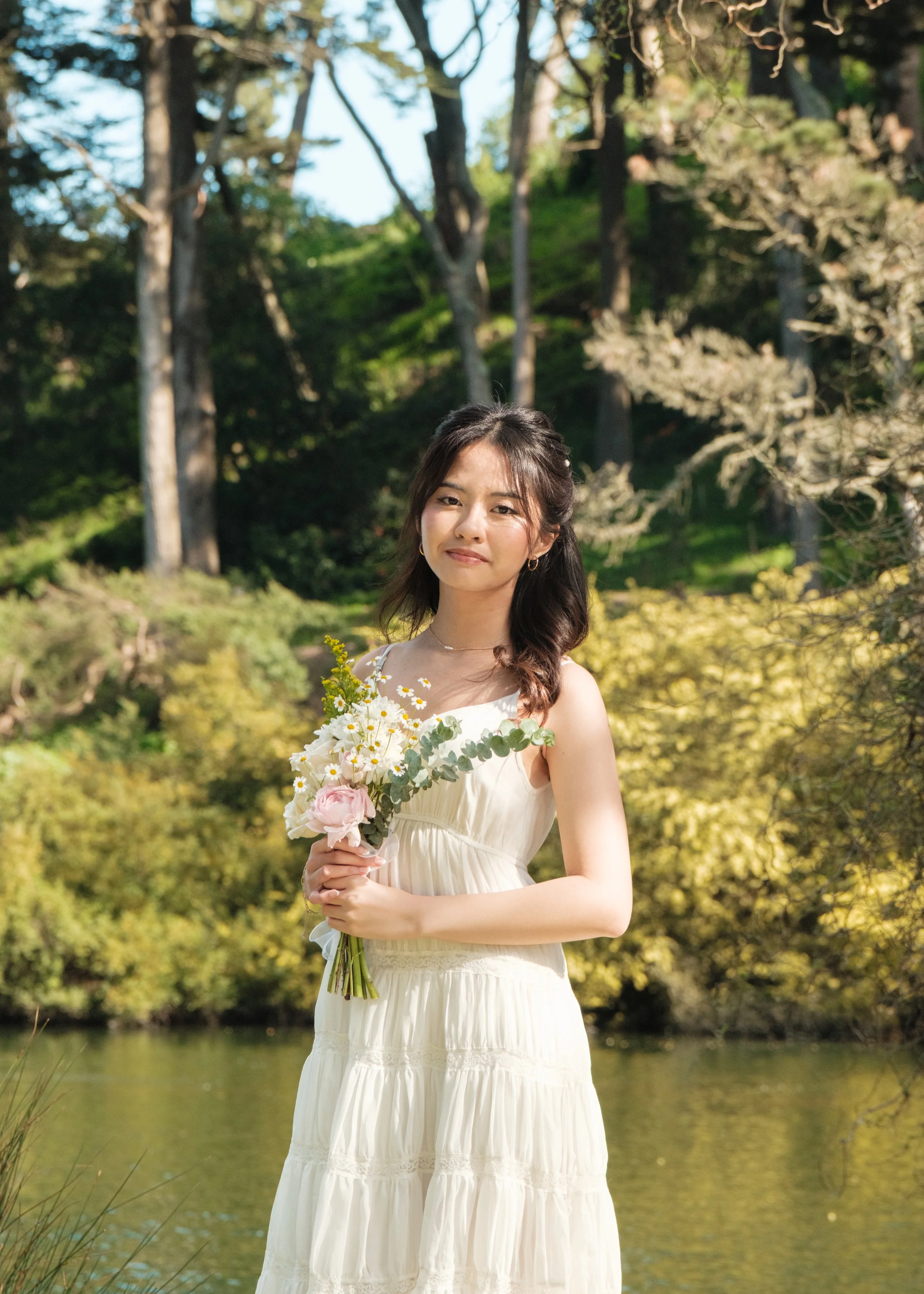 A woman in a white dress holding a bouquet of flowers, standing outdoors near a body of water with trees and greenery in the background.