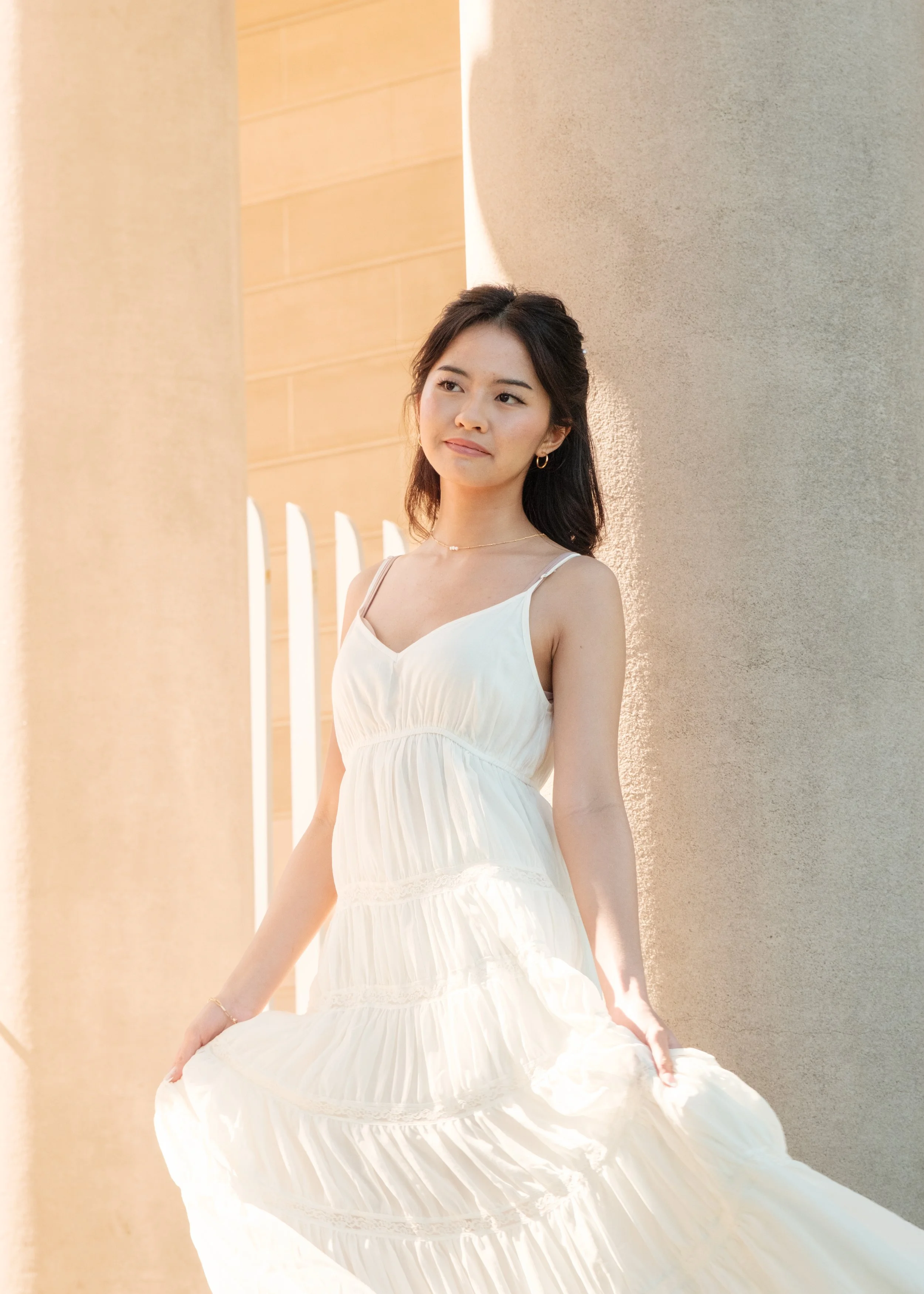 A young woman in a white dress standing outdoors near columns, softly illuminated by sunlight.