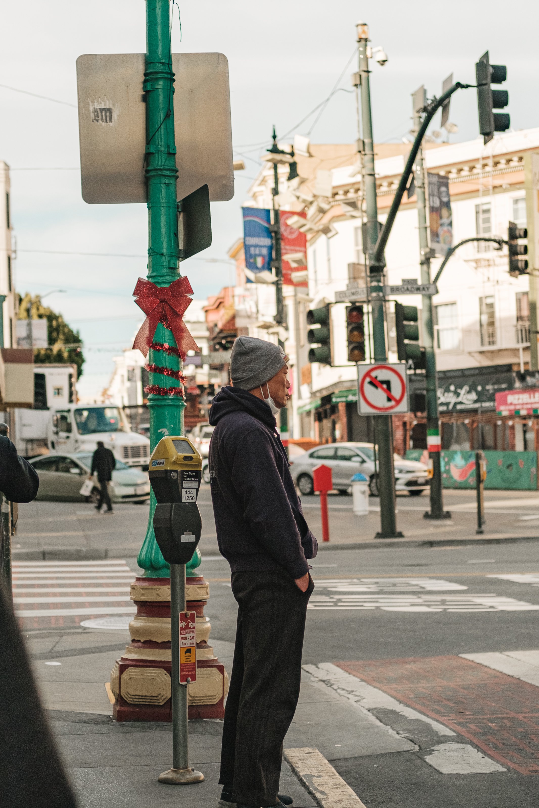 A man standing at a street corner with his hands in his pockets, wearing a gray beanie and a black hoodie, with a decorated green pole and busy city street in the background.