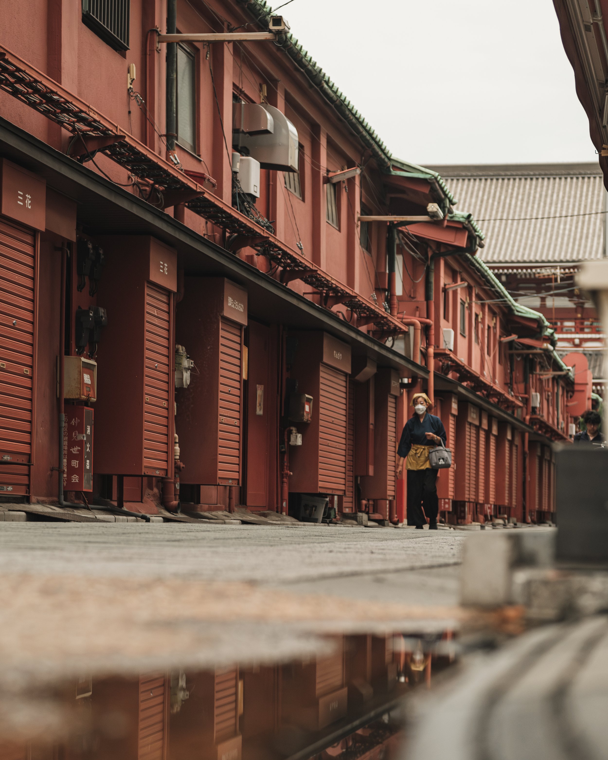 A woman wearing a face mask walks along a row of closed shop fronts in a traditional Japanese marketplace, with red buildings and green tiled roofs in the background.
