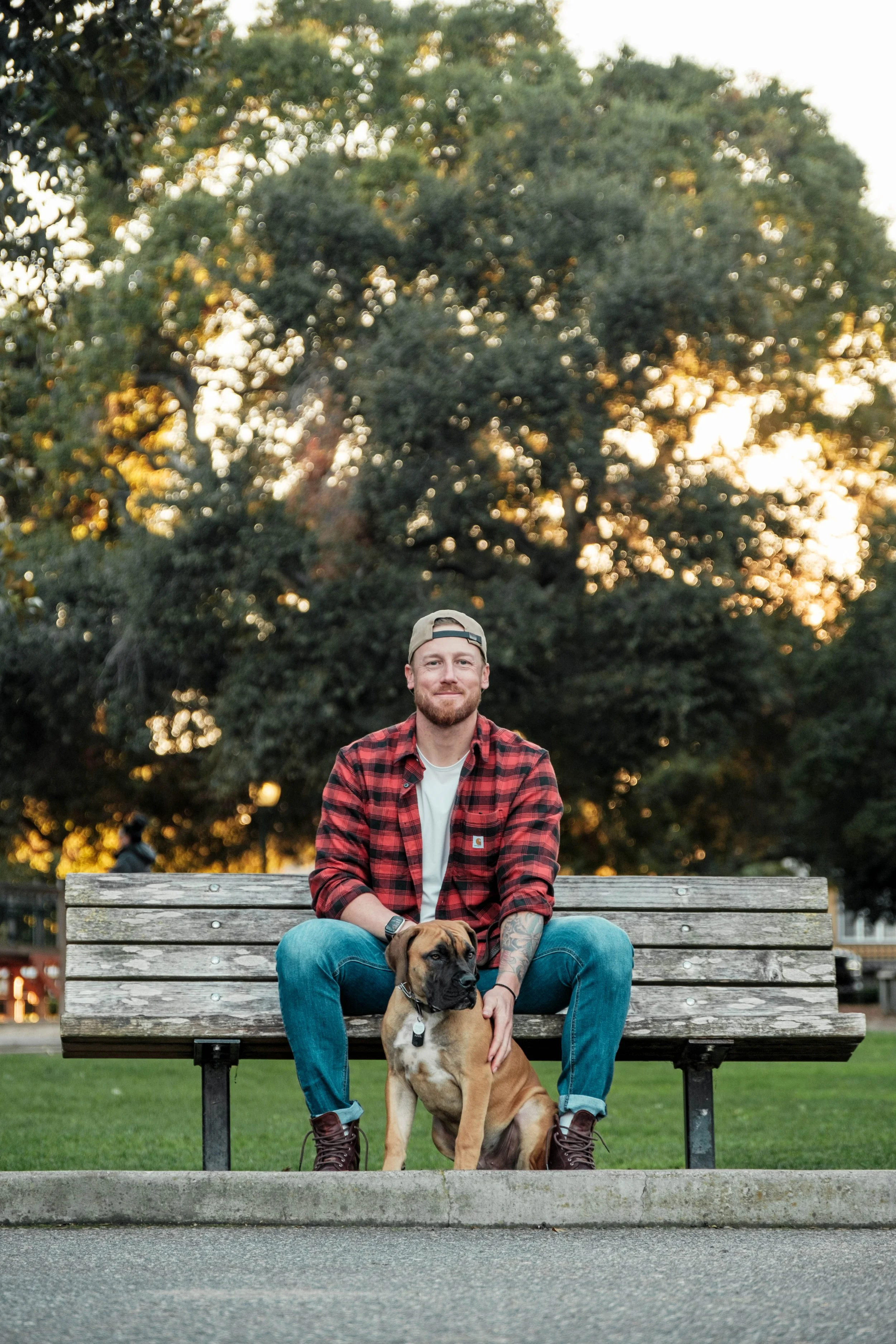 A bearded man wearing a red plaid shirt, white t-shirt, blue jeans, and a beige cap sitting on a park bench with a Boxer dog in front of him. They are outdoors with trees and a setting sun in the background.