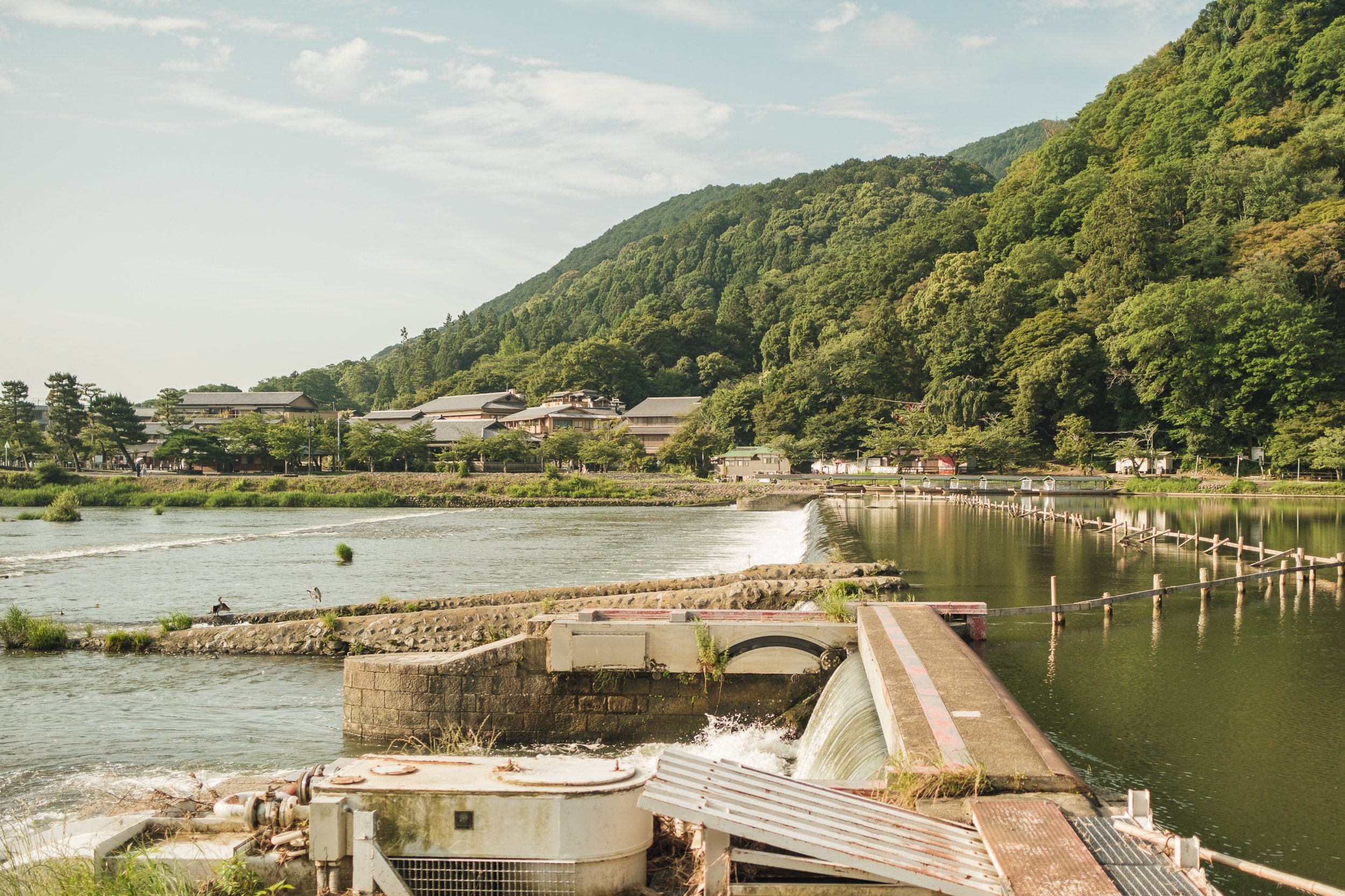 A scenic view of a river with a small waterfall, surrounded by green mountains and trees, with buildings in the background on a clear day.
