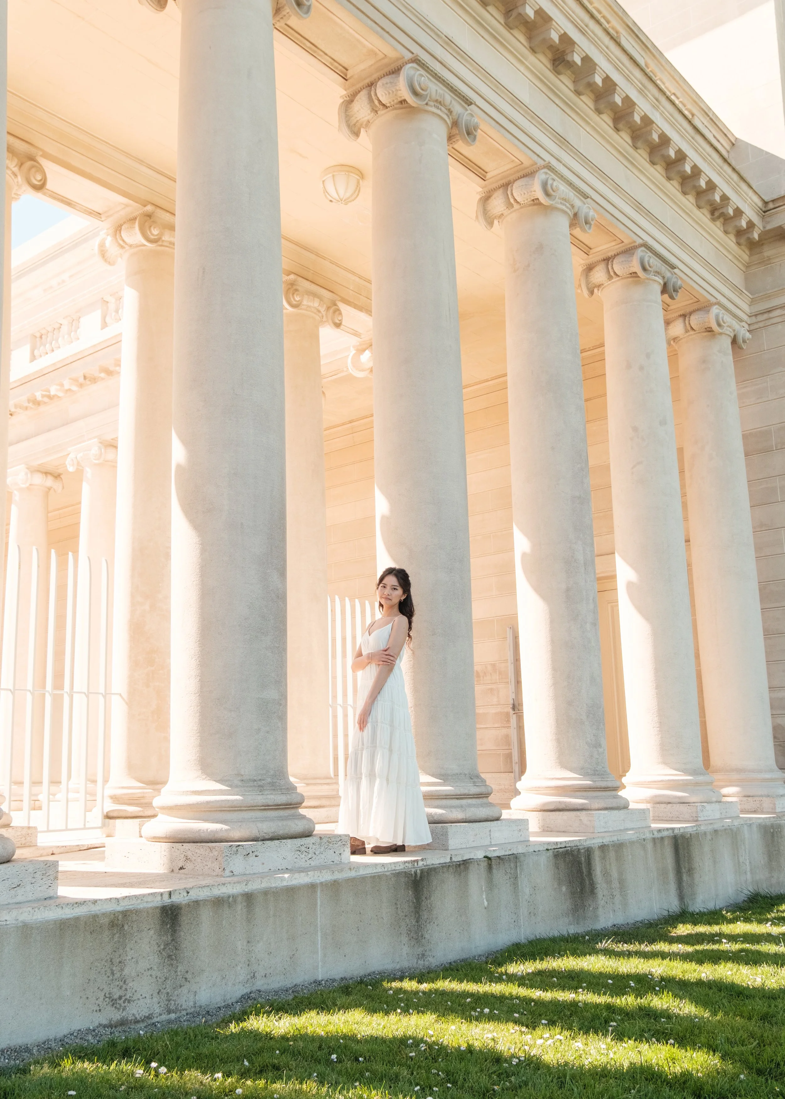 A woman in a white dress standing among large classical Greek-style columns outside a building during daytime.