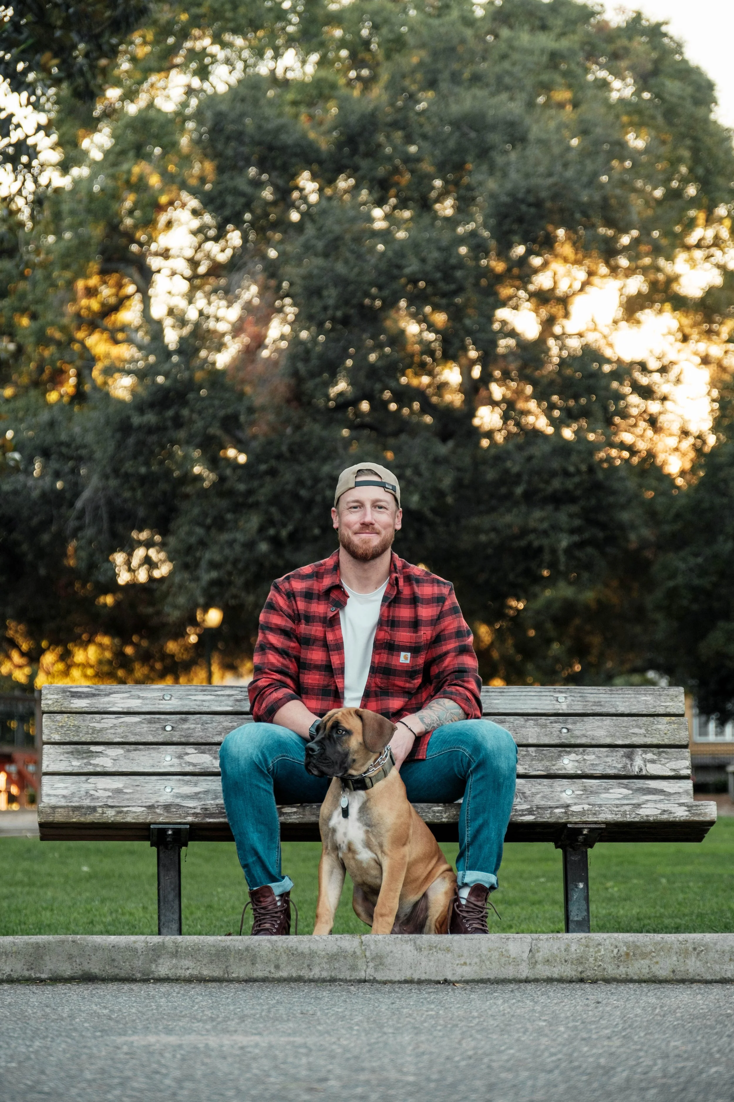 A man in a red plaid shirt and a beige cap sitting on a park bench with a brown dog. The man is smiling and the dog is sitting in front of him. There are trees and the sunset in the background.