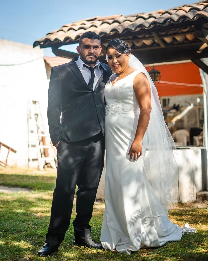 A bride and groom standing outdoors on a sunny day, dressed in wedding attire, with a rustic building in the background.