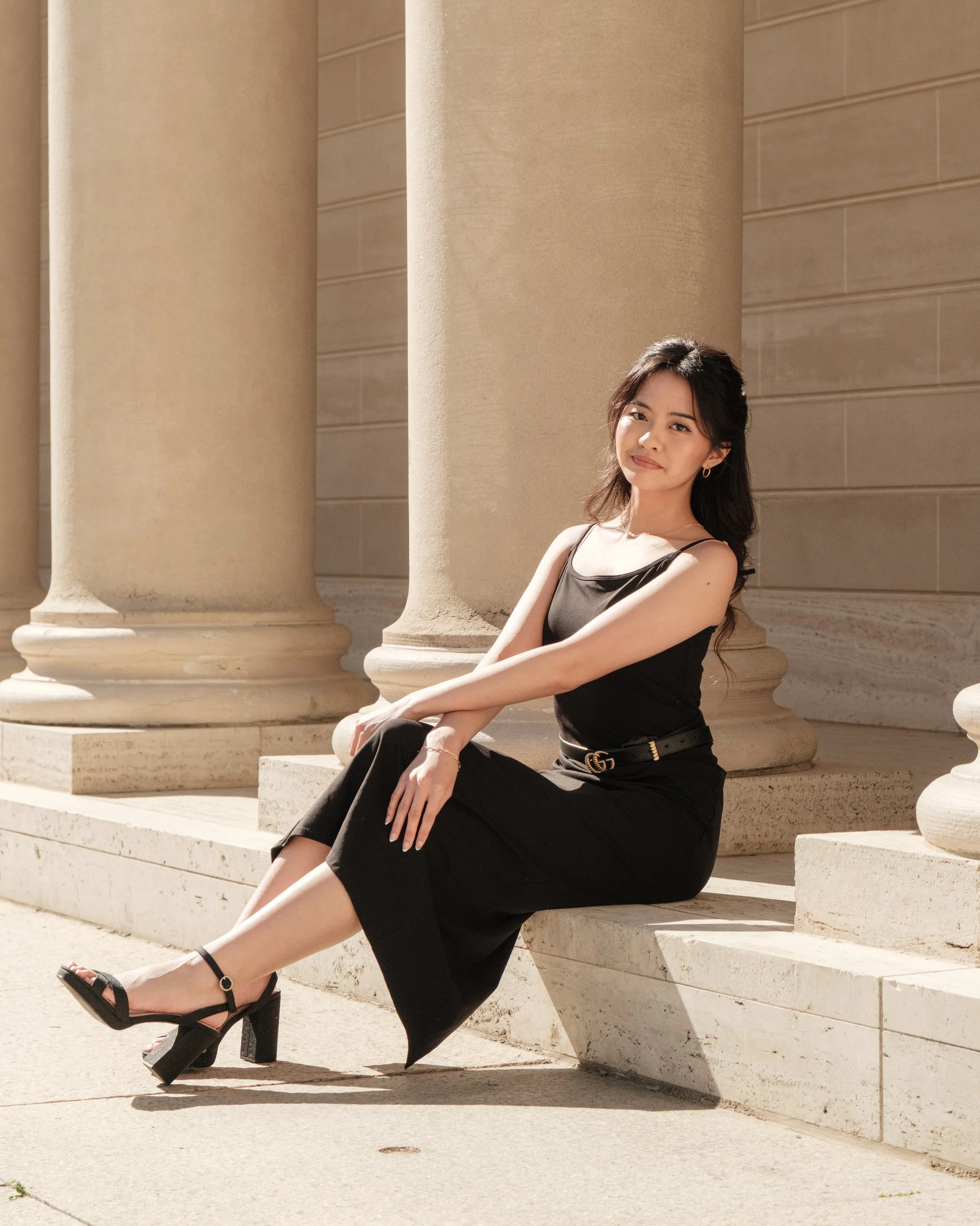 Young woman in black dress and high heels sitting on steps near large columns.