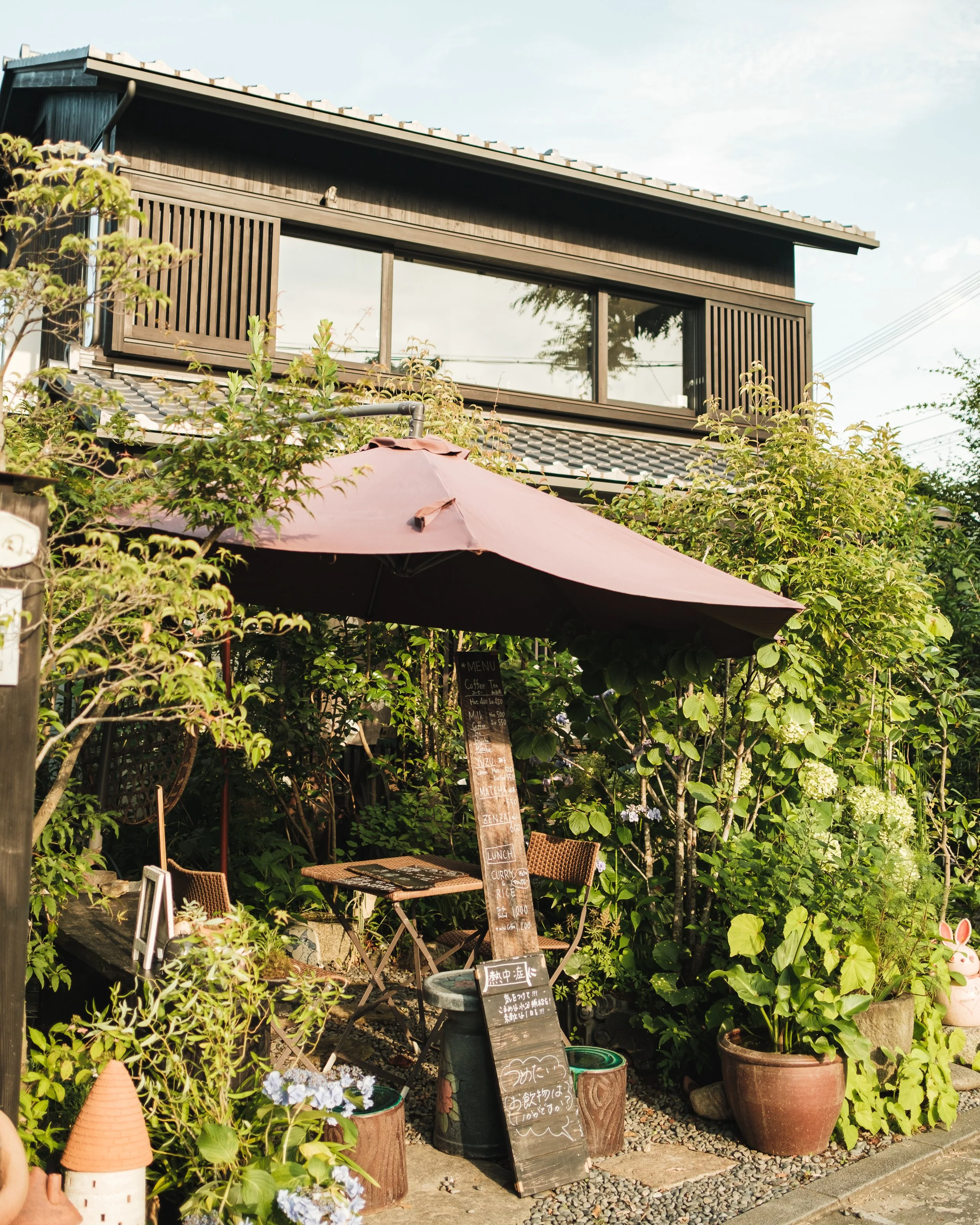 Outdoor cafe seating area with a large purple umbrella, surrounded by lush green plants, flowers, and potted plants, with a blackboard menu and a two-story wooden house in the background.